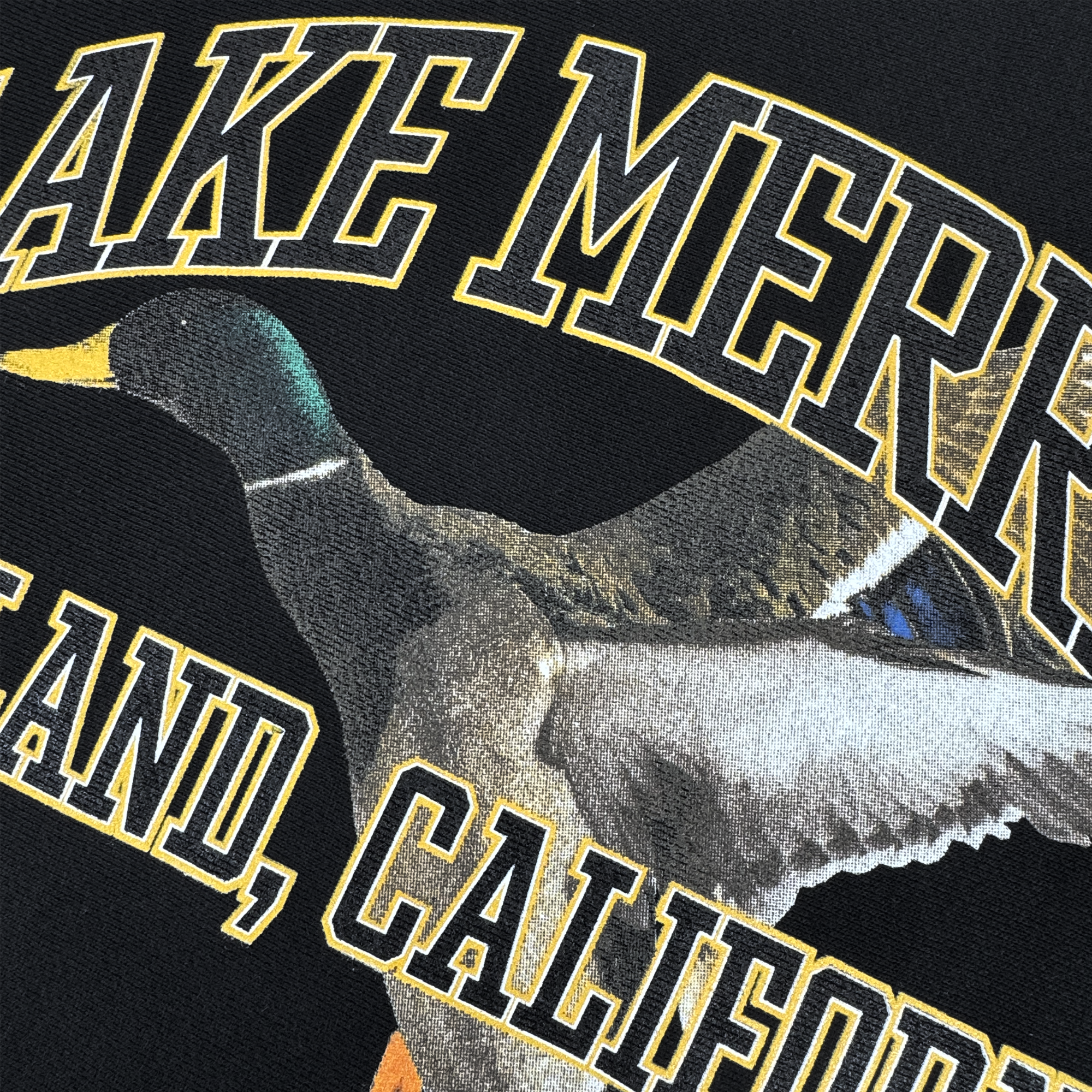 The Duck School Heavy Crew by Oaklandish features a male mallard duck in flight with "LAKE MERRITT" and "oakland, california" in bold yellow letters outlined in black. This striking design is available on a sleek black heavyweight fleece crewneck sweatshirt for men.