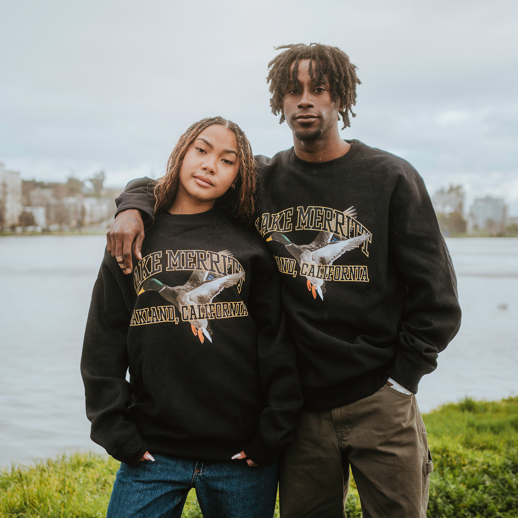 A person with long hair and another with short dreadlocks, both wearing Oaklandish Duck School Heavy Crew sweatshirts, stand by the lake. The one on the left has their arm around the other as they gaze into the camera, framed by trees and buildings.