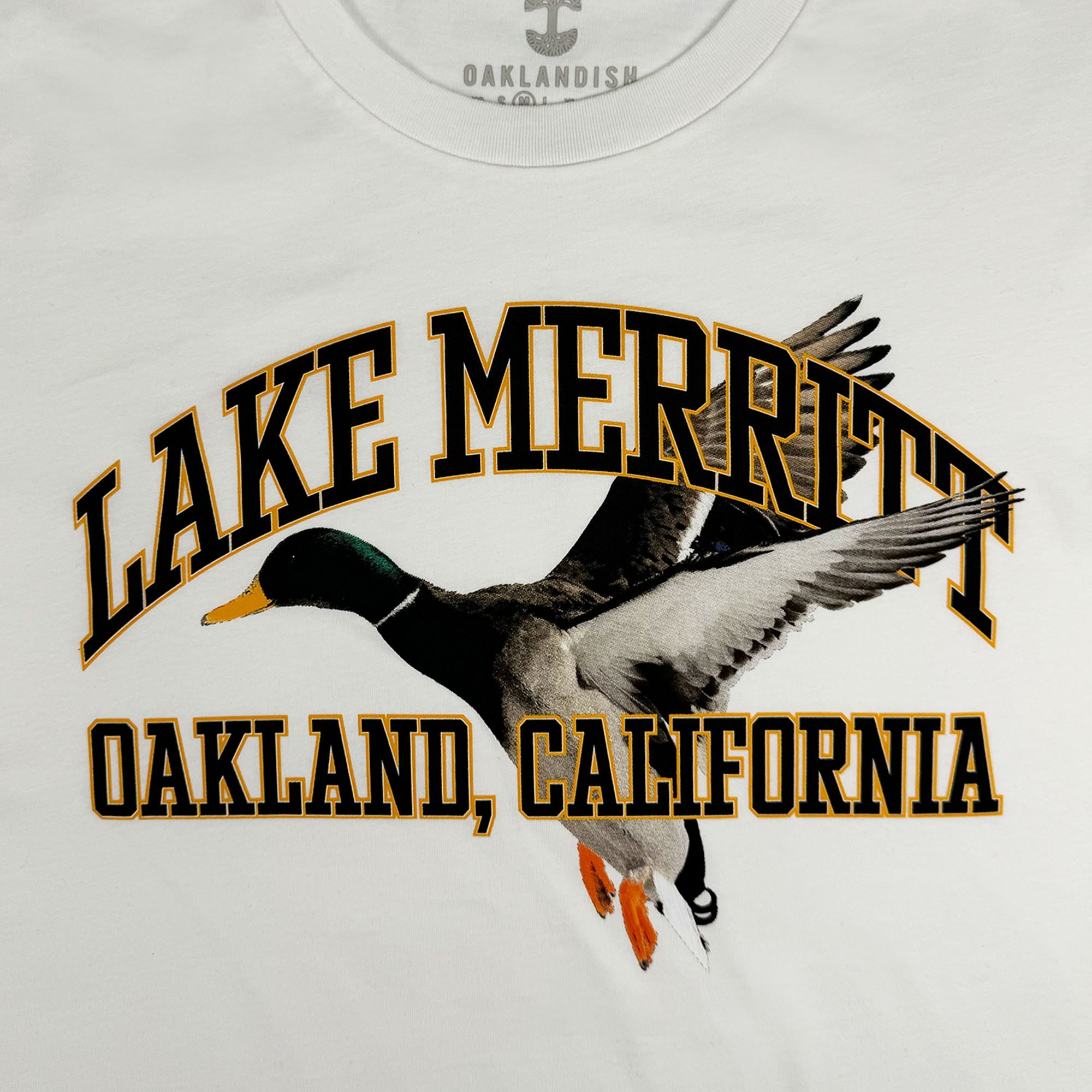 The image showcases the Duck School Tee by Oaklandish, a classic fit T-shirt made from 100% cotton. It features an illustration of a flying mallard duck, with "Lake Merritt" displayed in bold yellow and black above it and "Oakland, California" printed below.