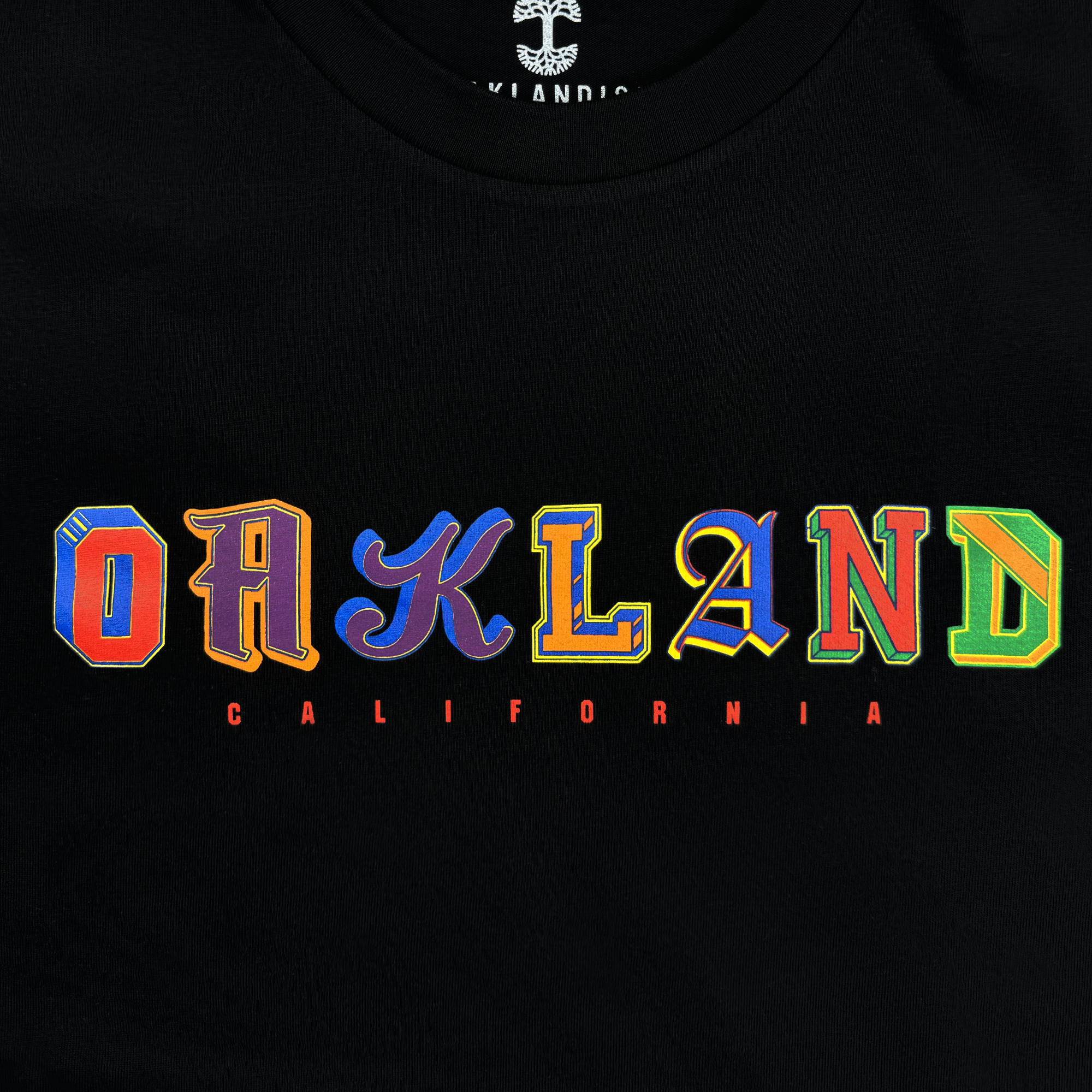 The image showcases the "First Team Tee" by Oaklandish, a black T-shirt made of 100% cotton. It features "OAKLAND" in vibrant, stylized lettering across the chest, with each letter having a unique design. Below this is "CALIFORNIA" printed in smaller red text. This classic fit tee effortlessly combines comfort and style.