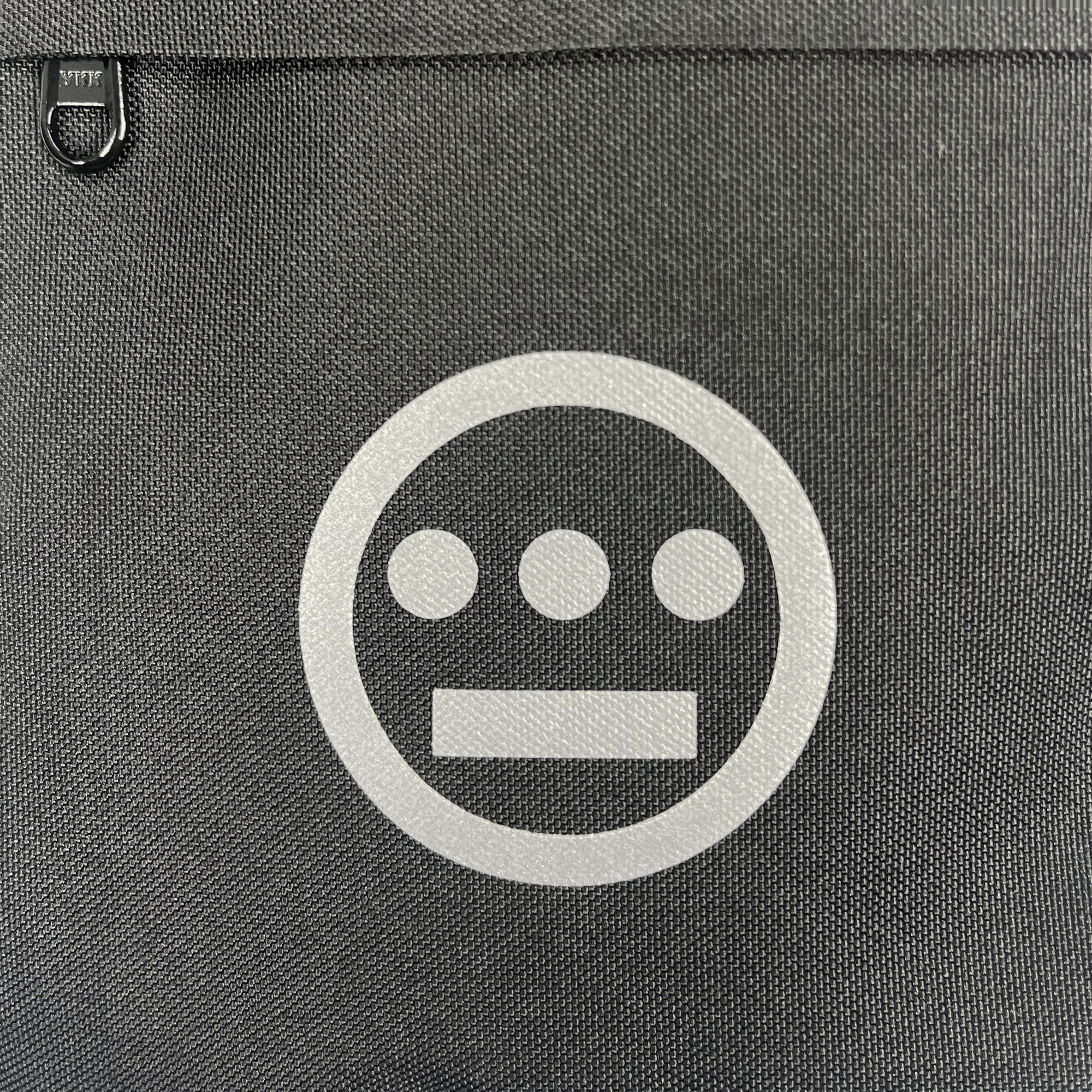 The Hiero Flight Bag by Hieroglyphics boasts a black textured surface with a minimalist white logo at the center, reminiscent of hieroglyphics. The logo consists of three filled circles in a horizontal row above a horizontal rectangle, all enclosed within a white circle. A small black D-ring near the top left corner adds an urban hip-hop crew vibe to the design.