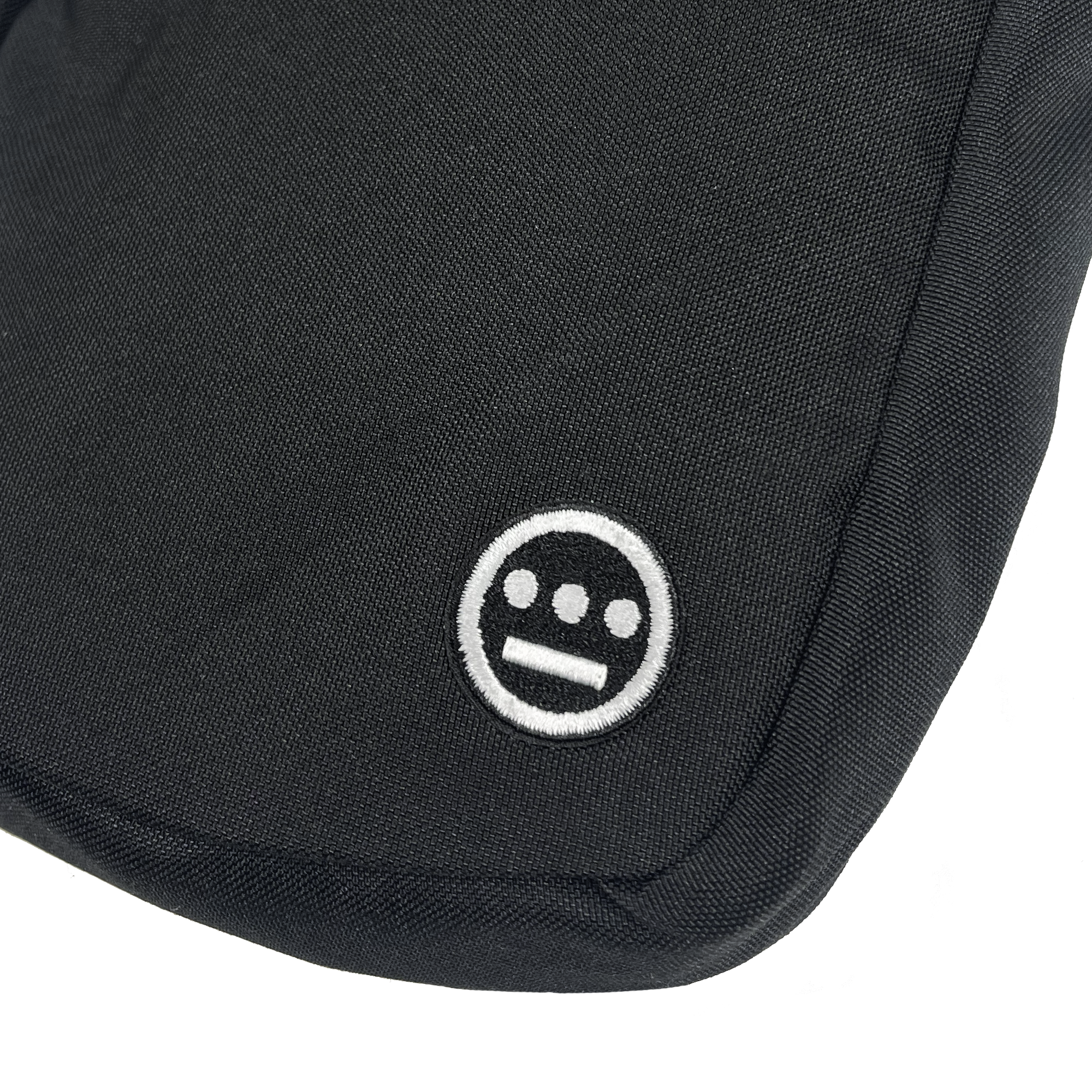 Close-up of the Hieroglyphics Hiero Flight Bag, a black polyester item, featuring a small white embroidery of the iconic Hieroglyphics symbol—a simple face with a straight line for a mouth and three dots for eyes and nose—at the bottom corner. The stitching detail and texture of the fabric are visible.