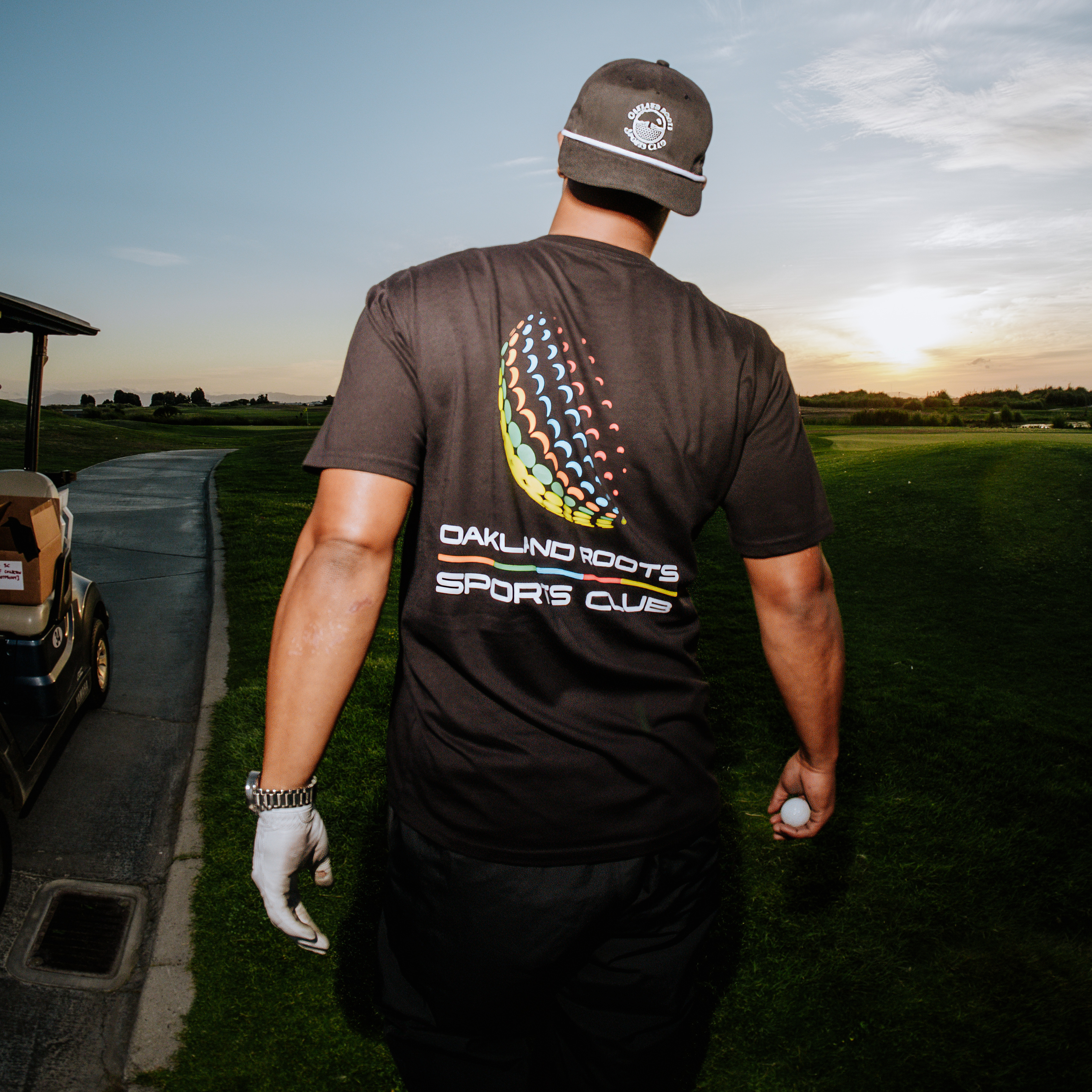 A man proudly wearing an Oakland Roots SC Foundations Golf Tee and matching cap leisurely walks along a golf course path at sunset. With a golf ball in one hand and a glove on the other, he embodies the spirit of social good. Nearby, a golf cart is parked as the sun sets behind partly cloudy skies.