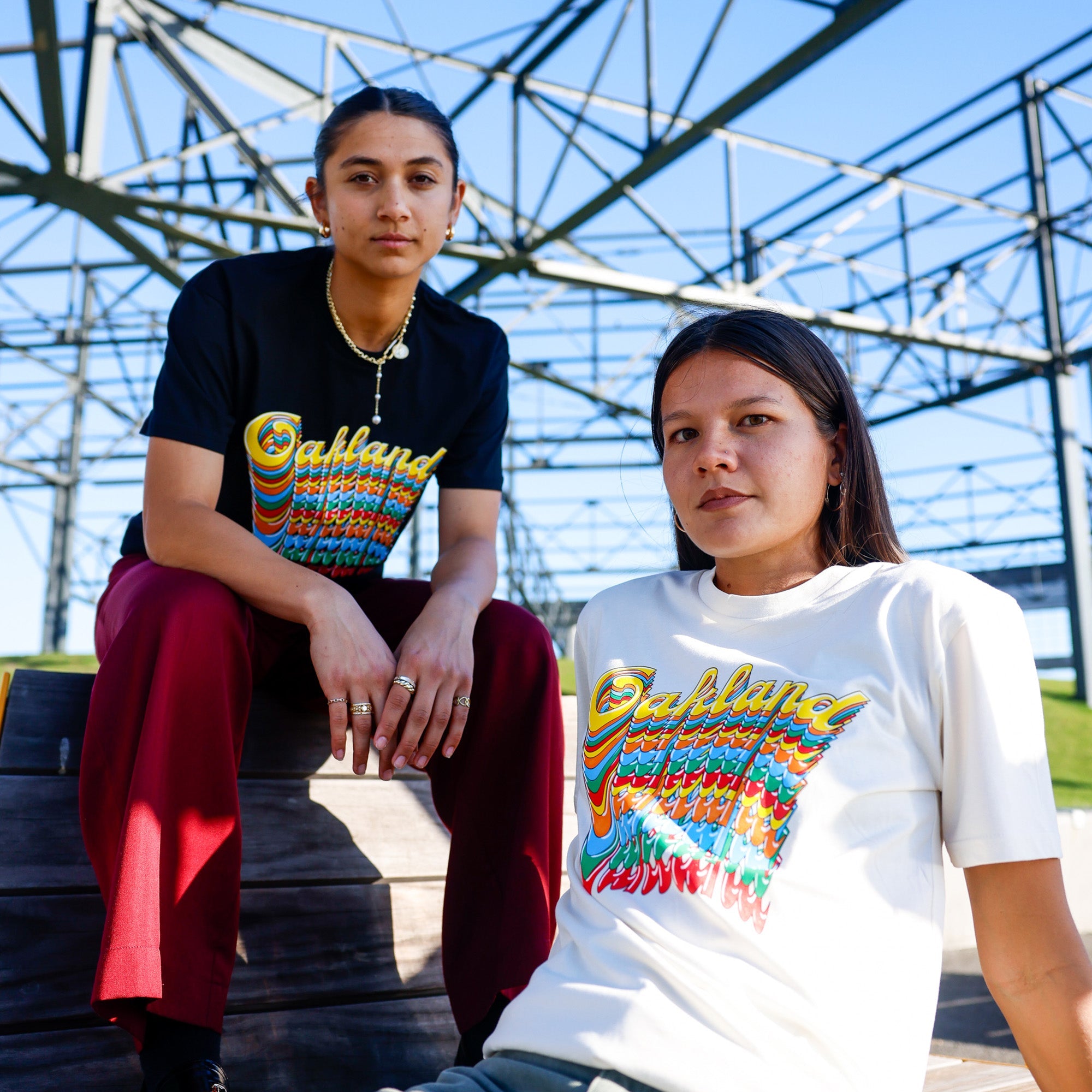 Two individuals are posing outdoors under a metal structure. One person, standing on the left, wears an Oakland Roots SC Funk Tee in black and red pants. The other person, sitting on the right, wears an Oakland Roots SC Funk Tee in white and green pants. Both shirts display a colorful "Oakland Roots" design. The sky is clear and blue.