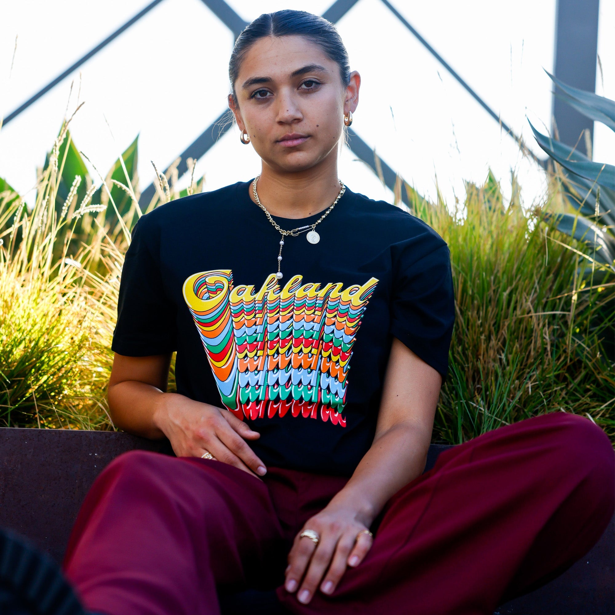 A person with long hair pulled back sits against a backdrop of lush green plants. They are wearing the Oakland Roots SC Funk Tee, a black T-shirt featuring a vibrant, wavy "Oakland Roots" graphic. Paired with maroon pants, multiple gold necklaces, and earrings, they have a serene expression while looking at the camera, embodying an essence of social good from the Oakland Roots SC brand.