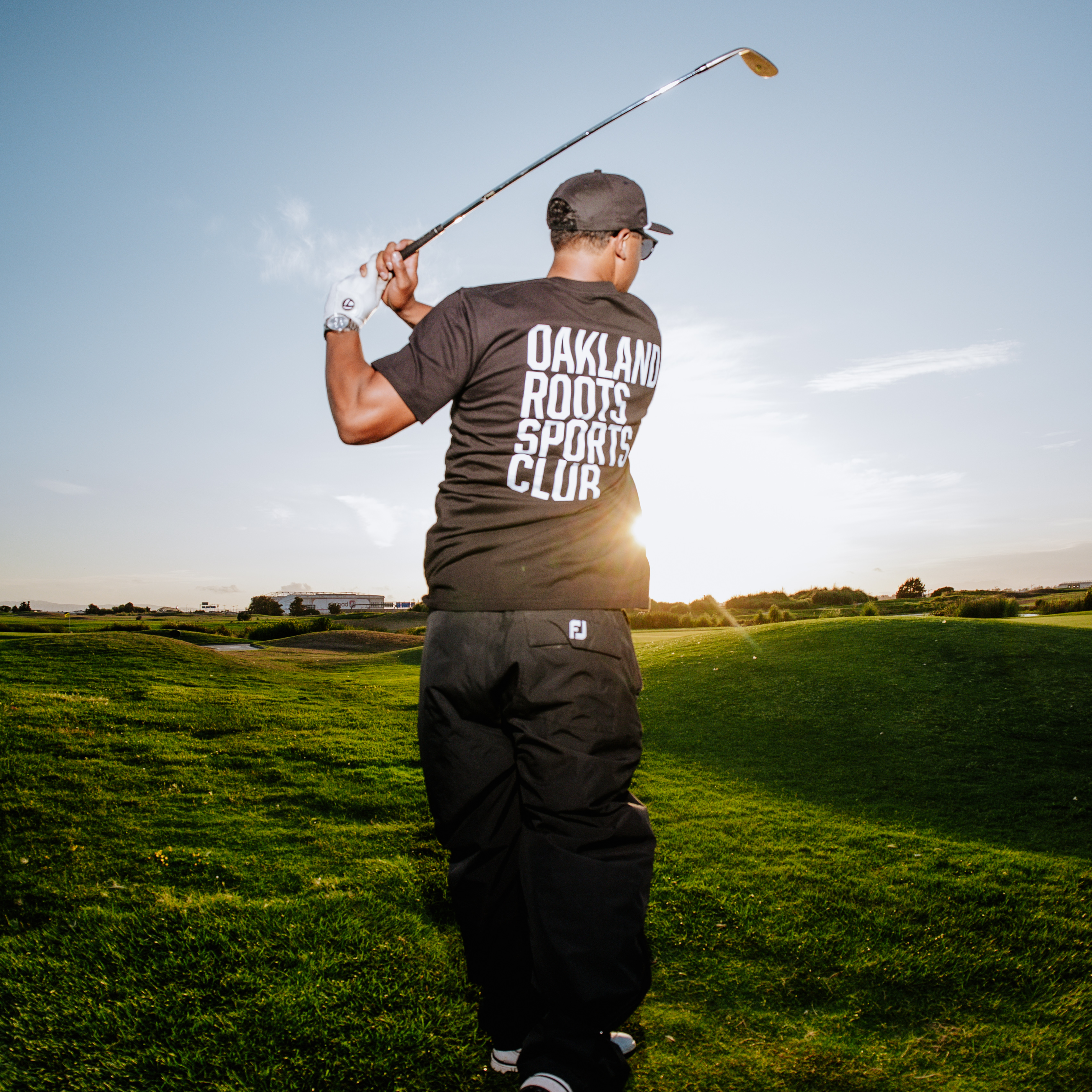 As the sun begins to set, a man immersed in the game of golf takes a swing on the green. He is dressed in black, wearing an "Oakland Roots SC" Golf Origins Tee that perfectly captures the essence of sports club camaraderie. The bright sun near the horizon casts long shadows across the field, allowing him to relish this moment of social enjoyment under a mostly clear sky with a few scattered clouds.