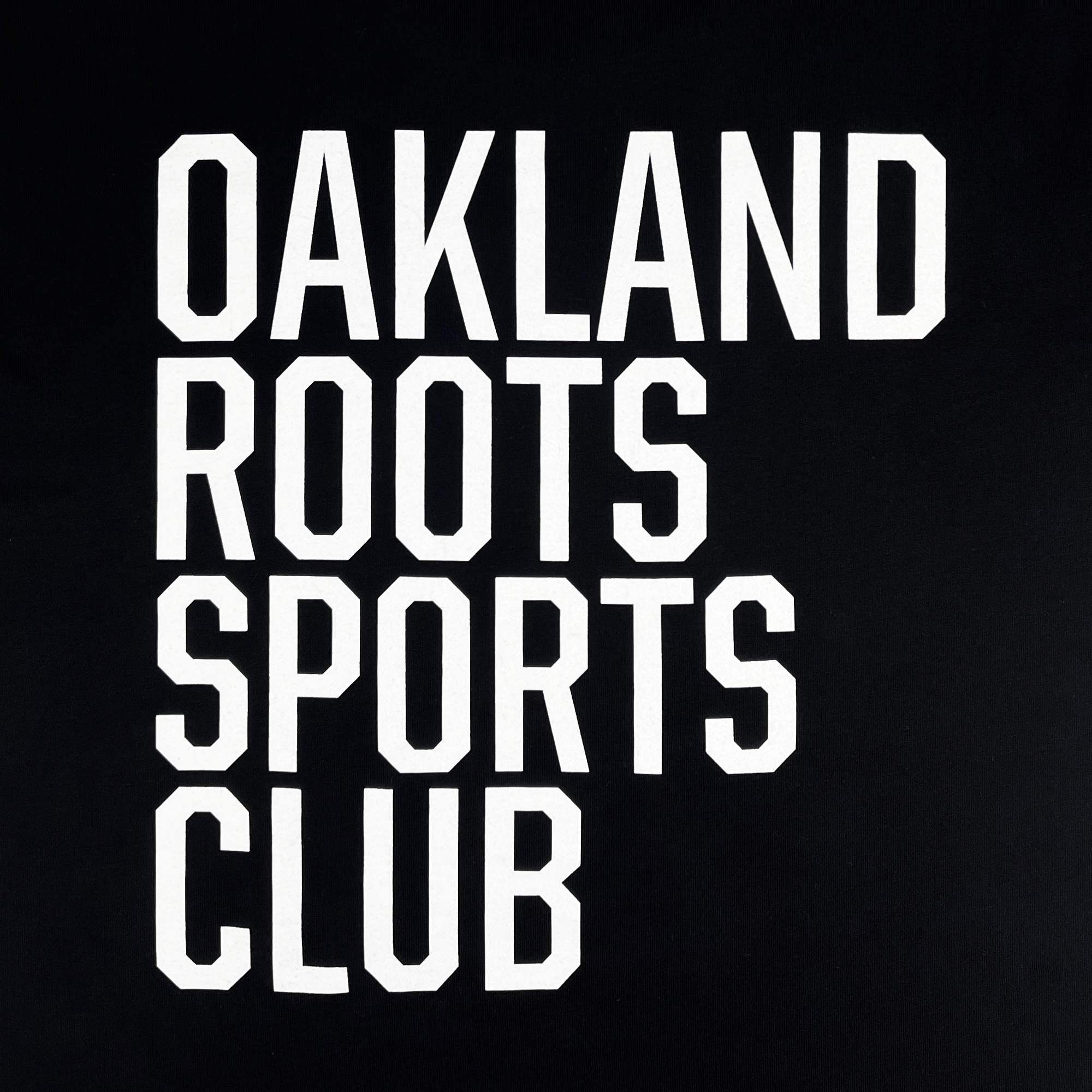 The image shows a black square adorned with bold white text in capital letters arranged across four lines. The text reads "OAKLAND ROOTS SPORTS CLUB." The thick, white font stands out starkly against the black background, capturing the essence of Oakland and the commitment embodied by Oakland Roots SC as showcased in their Golf Origins Tee.