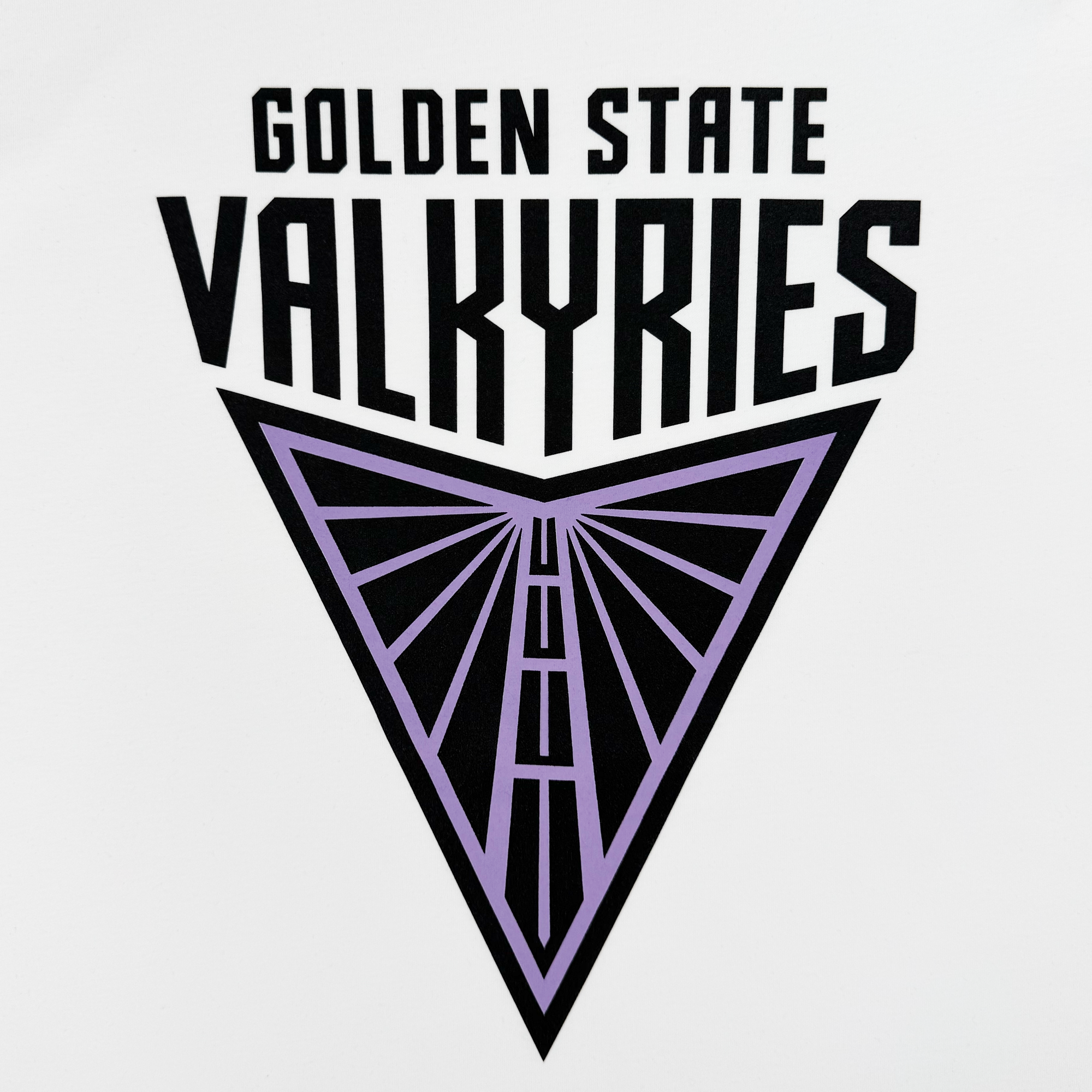 The image features the logo for "Golden State Valkyries" on a classic fit t-shirt named Golden State Valkyries Primary Tee by GSV. It boasts bold black text above an inverted triangle with a stylized purple wing outline, set against a crisp white 100% cotton backdrop.