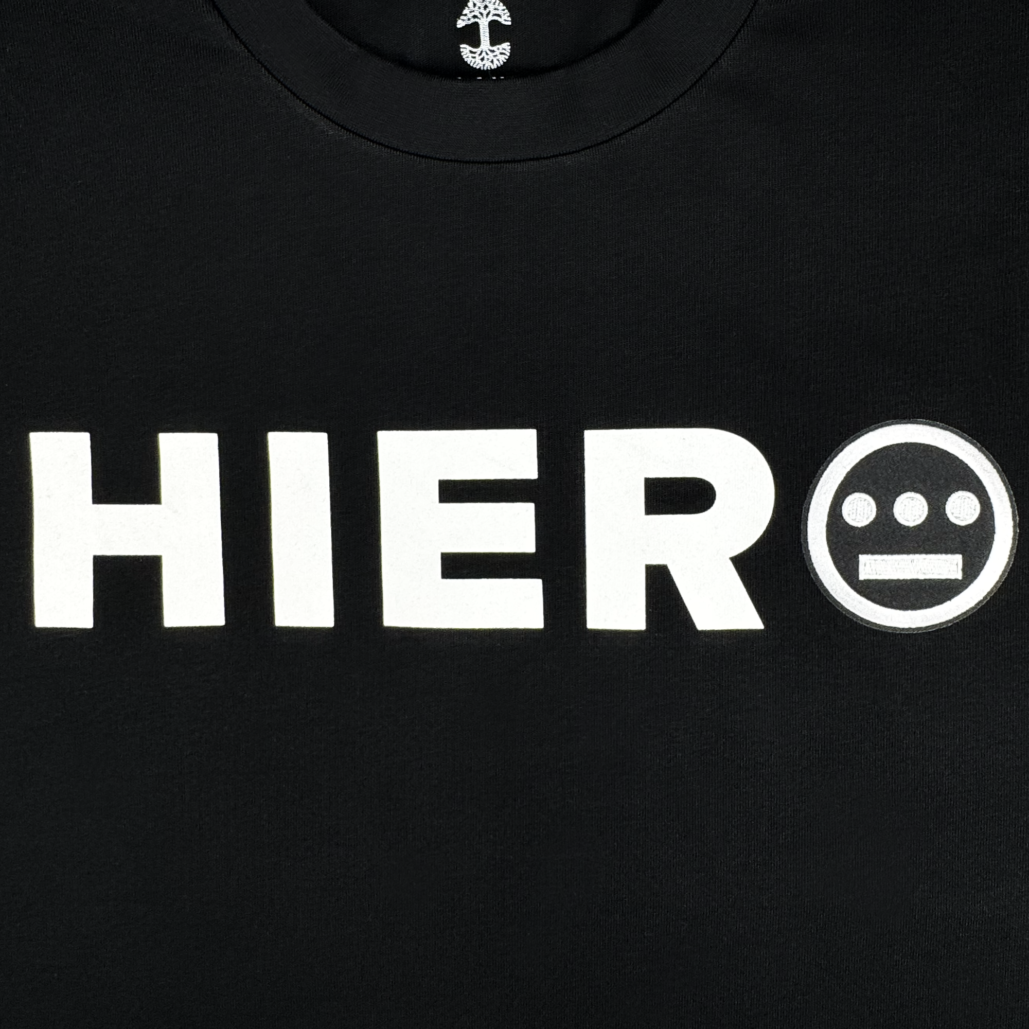 The Hieroglyphics Hiero Bold Heavy Tee in black features large white "HIER" text next to a circle logo with three dots over two, exuding a modern hieroglyphic style. A small white anchor at the neckline and smooth fabric infuse subtle hip-hop elements into its design.