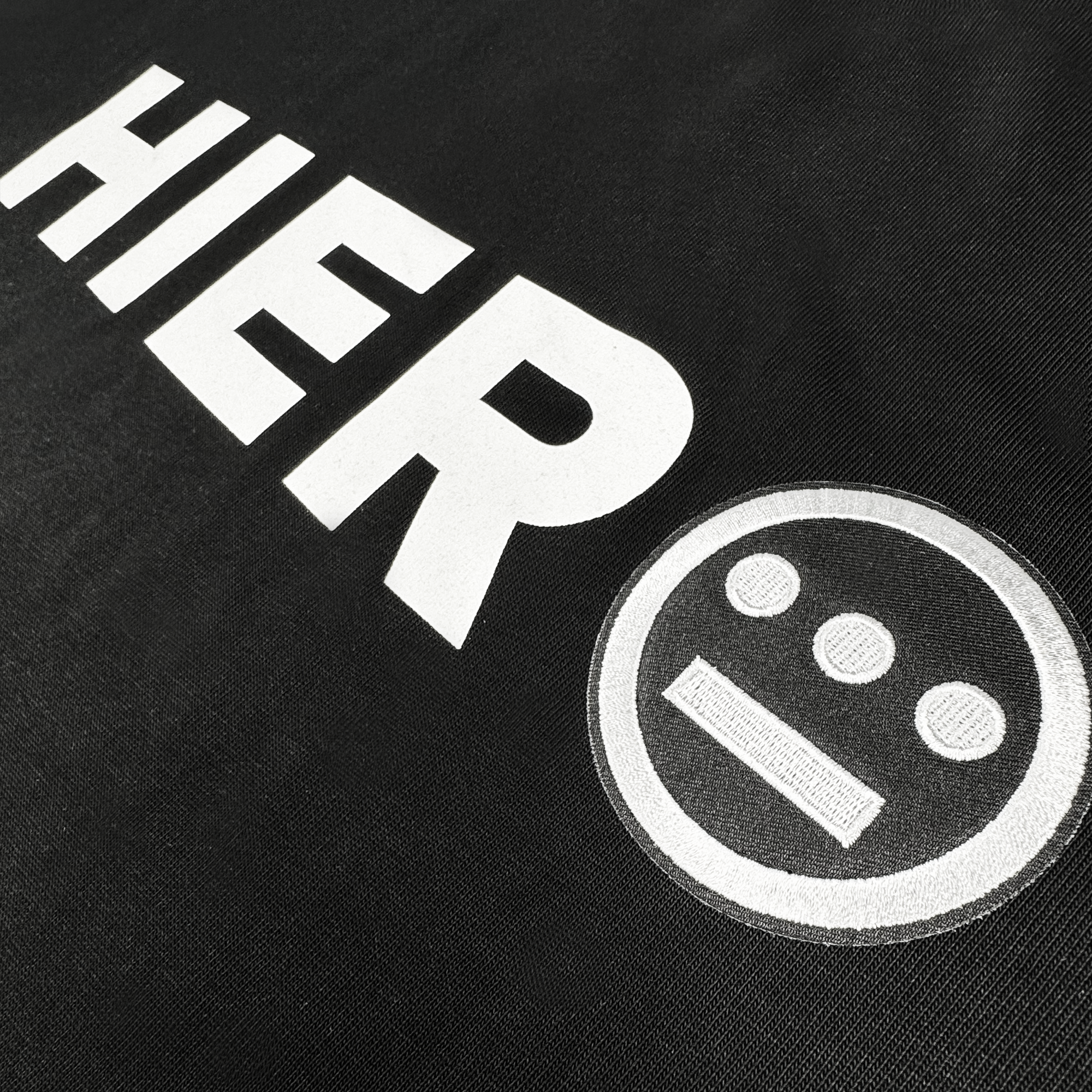 The Hiero Bold Heavy Tee by Hieroglyphics is a hip-hop inspired shirt with bold white "HIER" text and a circular hieroglyphic symbol, creating an eye-catching look on textured black fabric.