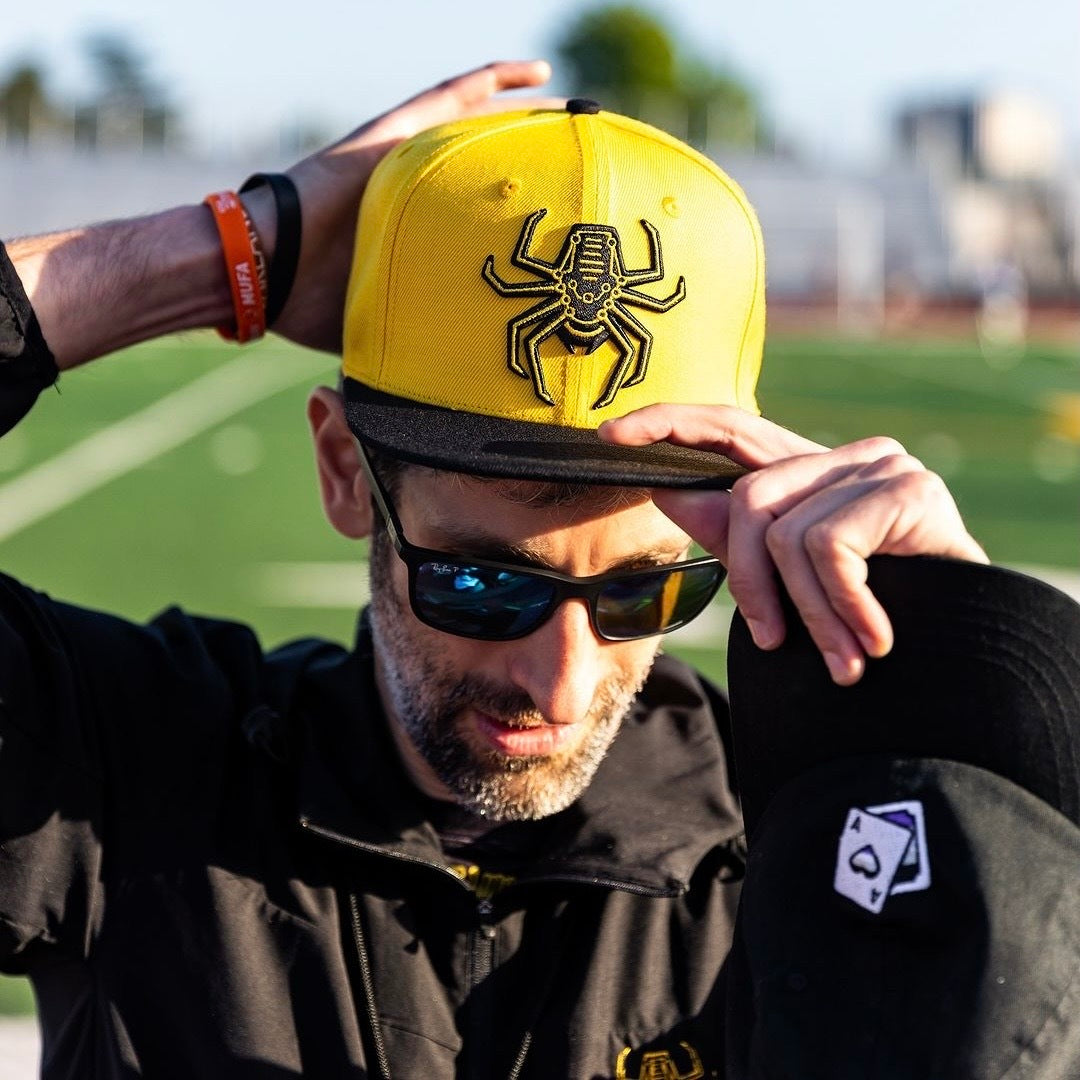 Guy with sunglasses on putting the yellow snapback on his head, outdoors.