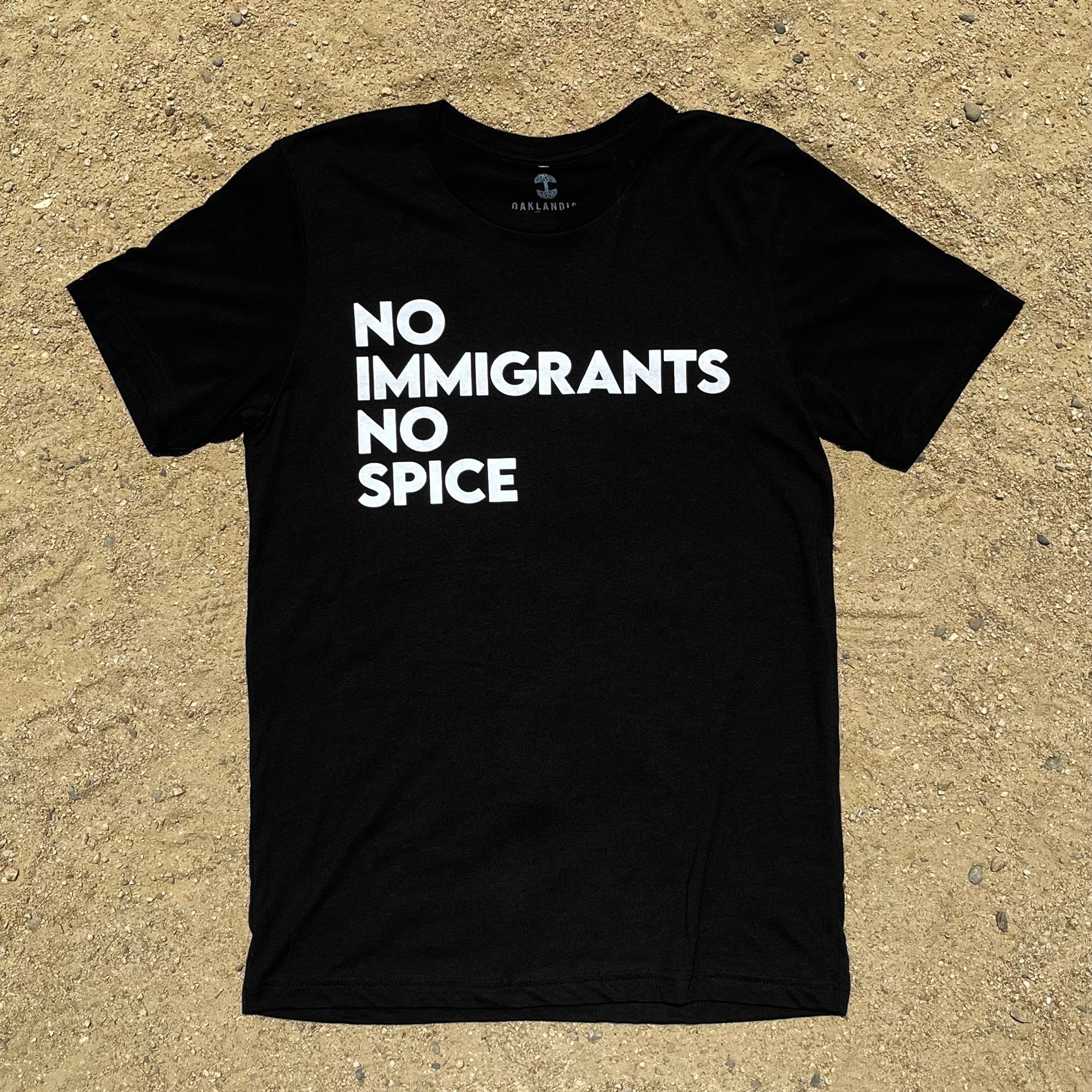 The NINS Classic Tee by Oaklandish, featuring bold white text "NO IMMIGRANTS NO SPICE," lies on a textured sandy surface, promoting #spicypride. This black T-shirt with short sleeves and a round neck is an essential statement piece.