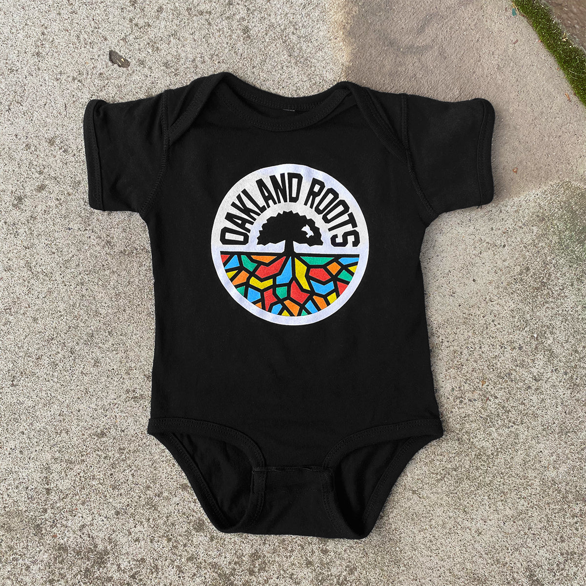 The Infant Oakland Roots SC Logo One-Piece, by Oakland Roots SC, is a black baby romper adorned with the Oakland Roots logo. It features a stylized tree above the words "Oakland Roots," with vibrant roots forming a mosaic of reds, greens, blues, and yellows below. This charming piece of sports club attire is displayed on a concrete surface.