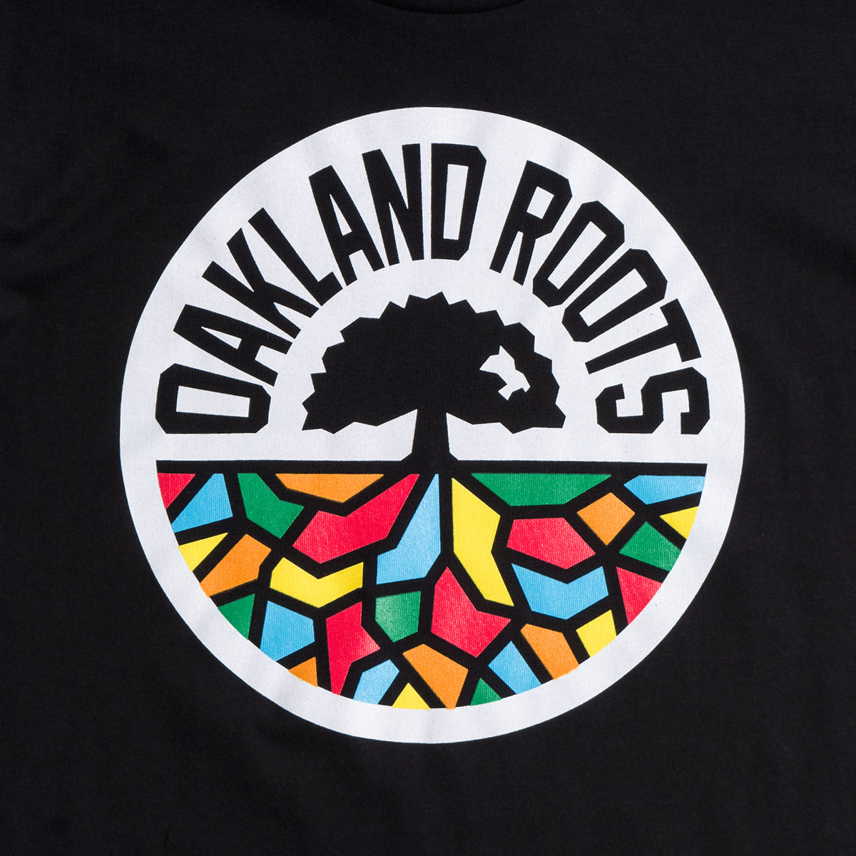 The Oakland Roots SC Classic Tee showcases a circular logo with the words "OAKLAND ROOTS" in bold black uppercase letters forming a semi-circle at the top. At the center of the logo, there is an illustration of a black tree with extended roots. Beneath the tree, reflecting their dedication to social good, is a vibrant mosaic background featuring sections in red, yellow, blue, and green.