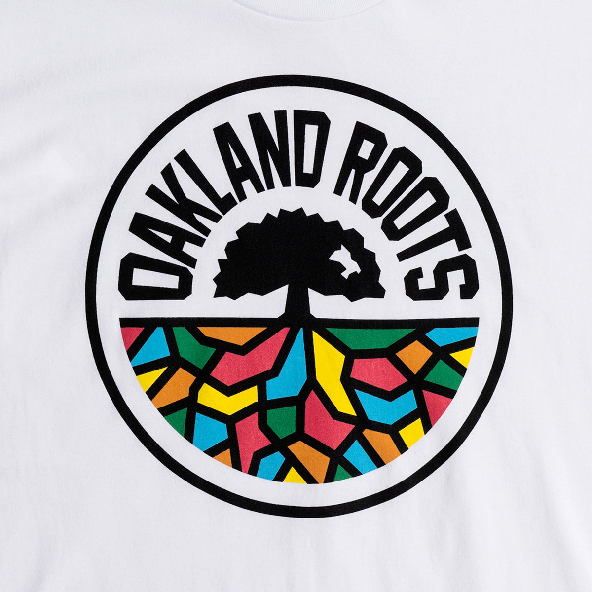 The image depicts the logo of Oakland Roots SC as featured on their Classic Tee. The logo showcases a black stylized tree with sprawling roots enclosed within a circular frame. Above the tree, the text "OAKLAND ROOTS" is arched. The lower half of the circle is adorned with a vibrant mosaic pattern in red, yellow, blue, and green, symbolizing their dedication to social good.