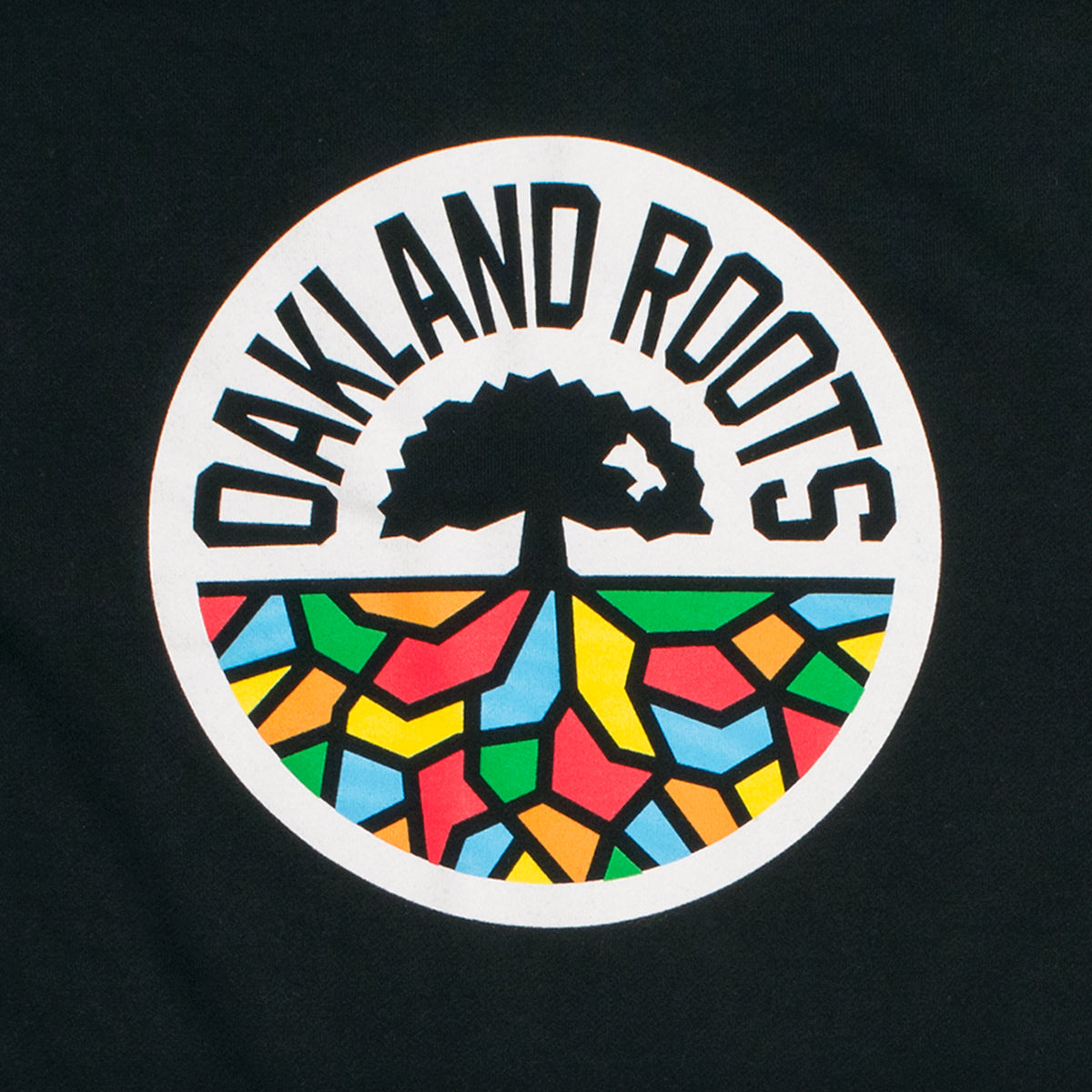 The image showcases the Oakland Roots SC Logo Crew by Oakland Roots SC, a sports club celebrated for its dedication to social good. The logo features a black tree with roots extending into colorful, abstract geometric shapes. This intricate design is encased within a white circle, with "OAKLAND ROOTS" in bold black letters curving around the upper half against a black background.