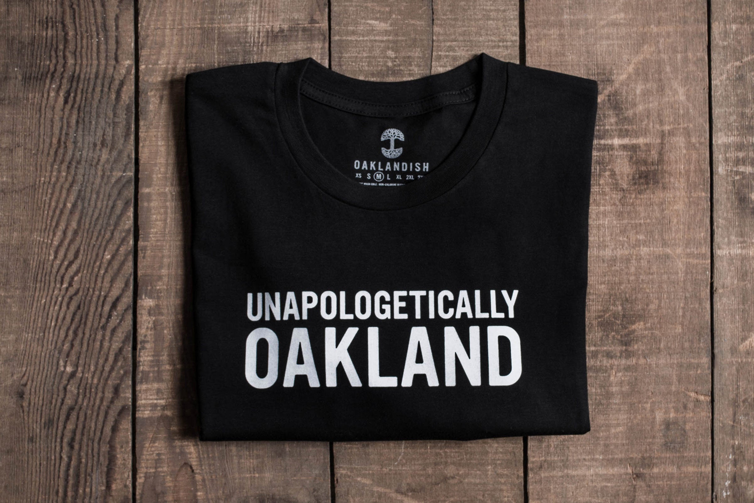 A folded black men's T-shirt, named "Unapologetic Tee," made from 100% cotton, displays white text "UNAPOLOGETICALLY OAKLAND" on a wooden surface. The tag shows the Oaklandish logo with faint branding details visible.