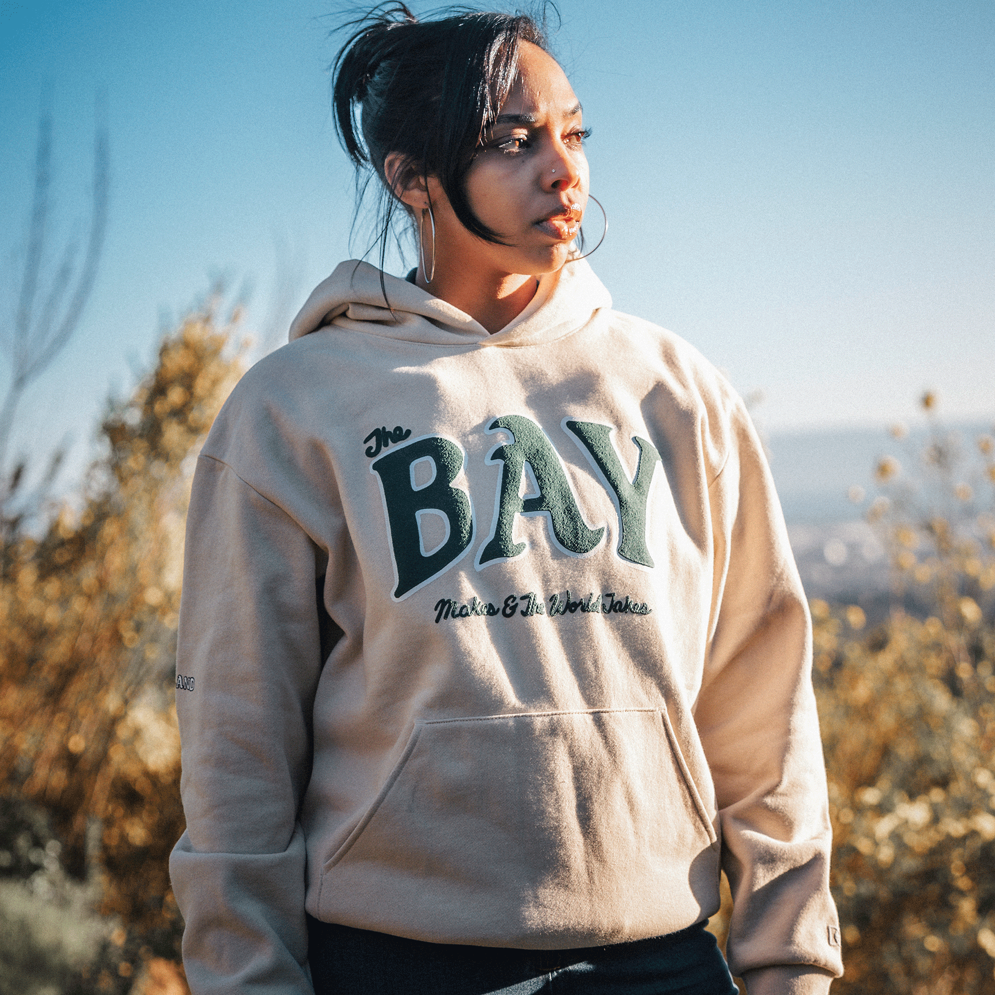 A young woman stands outdoors, her face illuminated by the sunlight. She is wearing The Bay Hoodie by DOC from Oaklandish's capsule collection, which features "The BAY" and "Make Of The World Yours" printed on premium heavyweight fleece. The background shows a blurred outdoor setting with shrubs and a clear blue sky, indicating a sunny day.