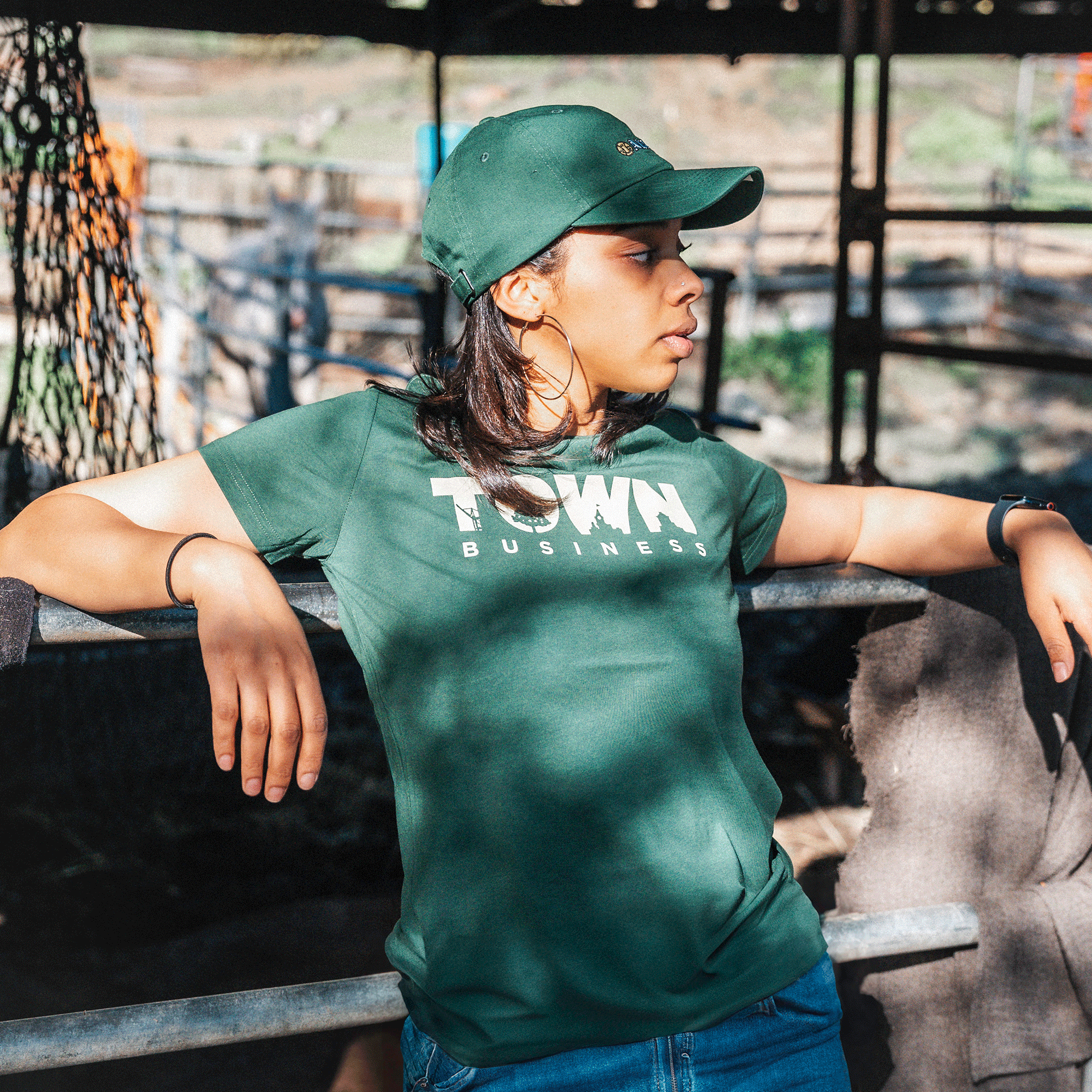 A person in a green cap and an "Other" brand Women's Town Business 2024 Tee leans casually on a metal railing. The East Bay-inspired backdrop with wooden fencing and blurred foliage captures their relaxed mood as they enjoy the sunny day in Oakland.