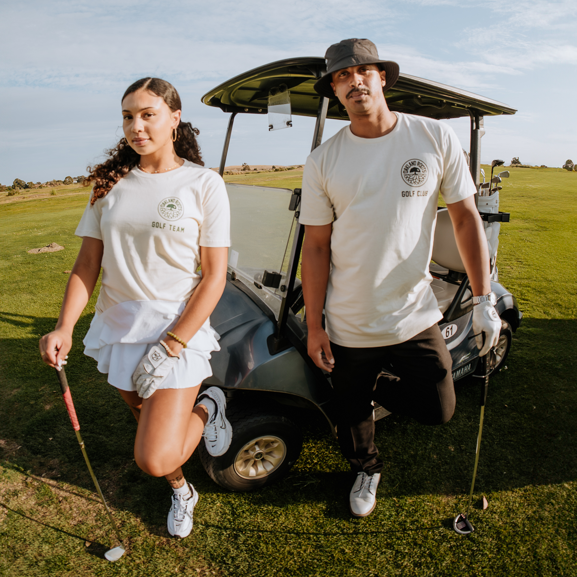 A man and a woman stand confidently in front of a golf cart on a lush golf course, embodying the unity and spirit of Oakland Roots SC. Both are dressed in white golf attire, with the woman wearing a skirt and the man in shorts. They each hold golf clubs and wear gloves, ready to play under the clear sky with a light breeze. The man proudly sports an Oakland Roots SC Members Only Tee.