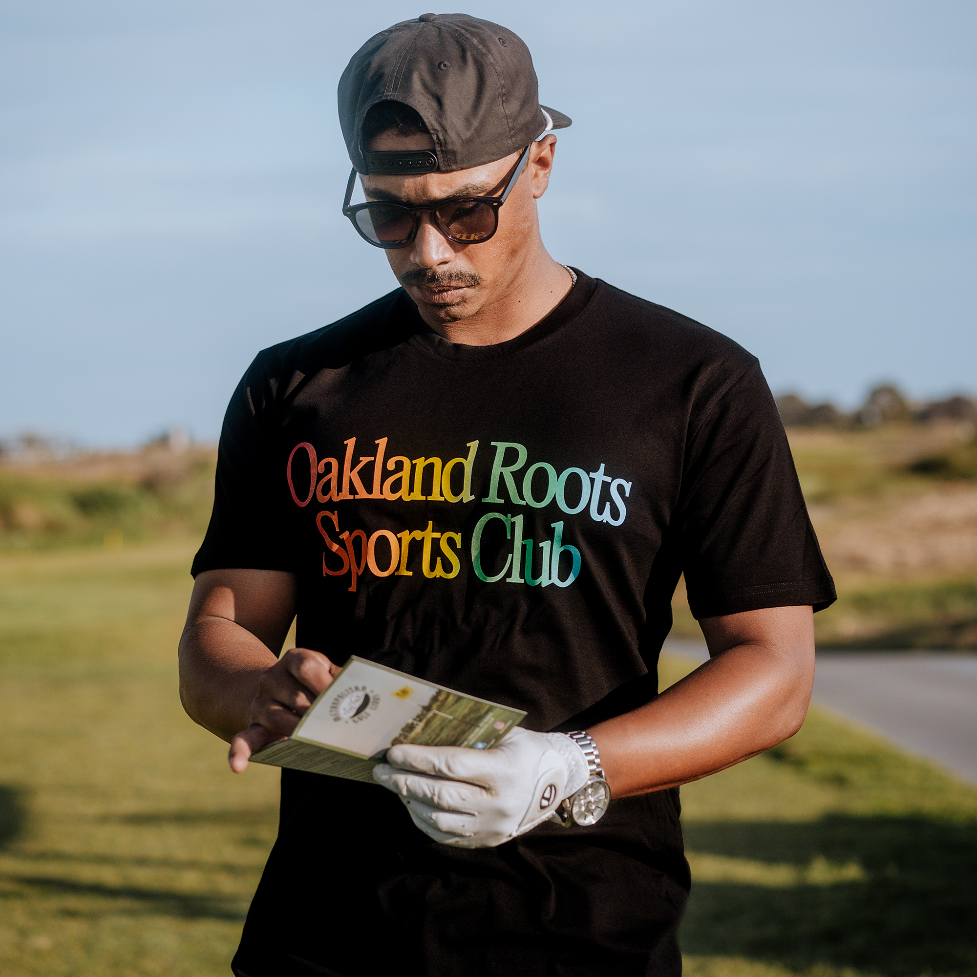 A man stands outdoors on a sunny day, wearing a backward baseball cap, sunglasses, and a "More Than Soccer Tee" from Oakland Roots SC that promotes social good. He is looking at an open booklet in his hands and has a white glove on one hand. The background shows a grassy area and a pathway.