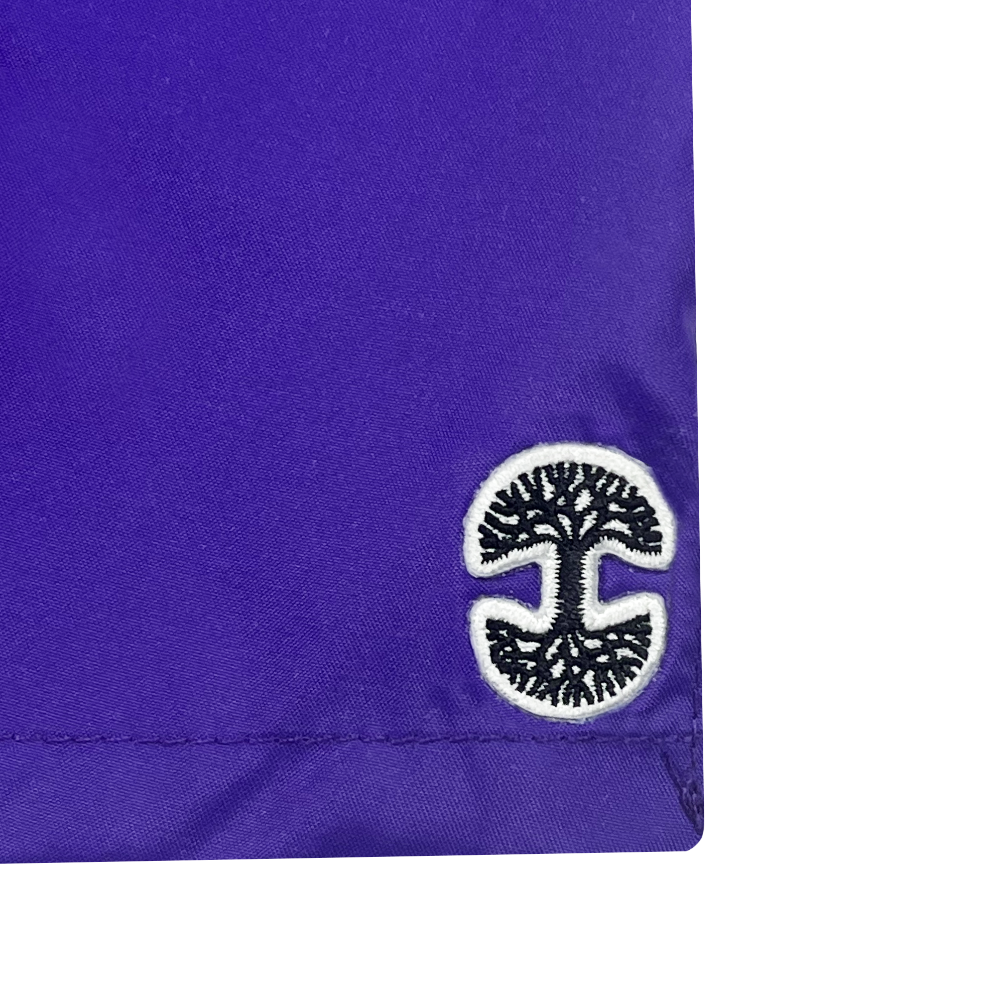 A close-up of the Women's Bandit Nylon Short by Oaklandish in purple fabric, featuring a black and white embroidered logo of a stylized tree with spreading branches and roots. The stitching around the hem of the short is visible against a plain white background.