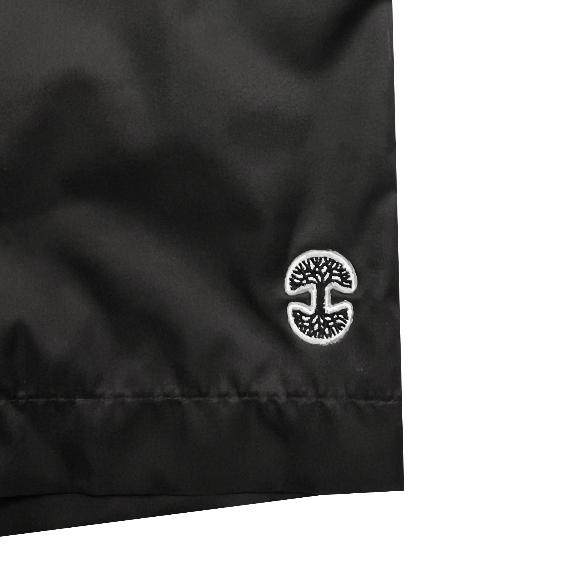 Close-up of a black fabric with an embroidered white and black circular logo featuring a stylized tree. The bottom hem of the fabric is visible, suggesting it's part of Oaklandish's Bandit Nylon Short. The smooth, slightly shiny texture adds to its sleek appearance.