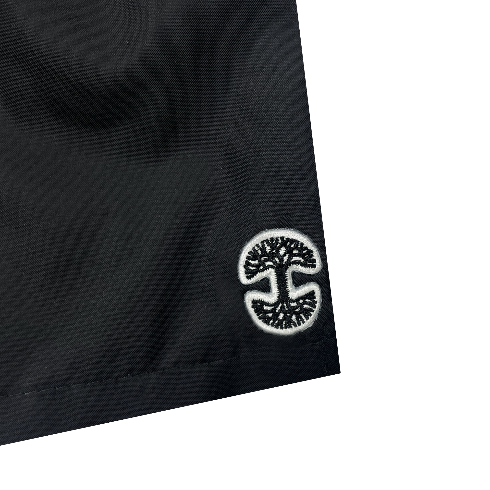 Close-up of the corner of a black fabric item, likely Oaklandish Women's Bandit Nylon Shorts. The edge is neatly sewn, and near the corner, there is an embroidered white circular logo featuring a stylized tree with branches and roots mirrored symmetrically. The background is plain white.