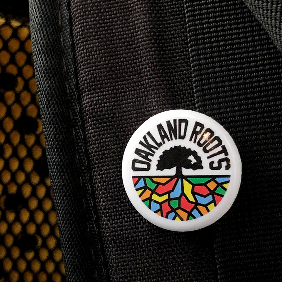 The Oakland Roots SC Pin, a circular badge, is attached to a black woven fabric and proudly displays the "OAKLAND ROOTS" logo. This logo symbolizes a sports club dedicated to social good, showcasing a stylized black tree with roots expanding into a vibrant mosaic pattern at the base. The words "OAKLAND ROOTS" are boldly written in uppercase letters around the top edge.