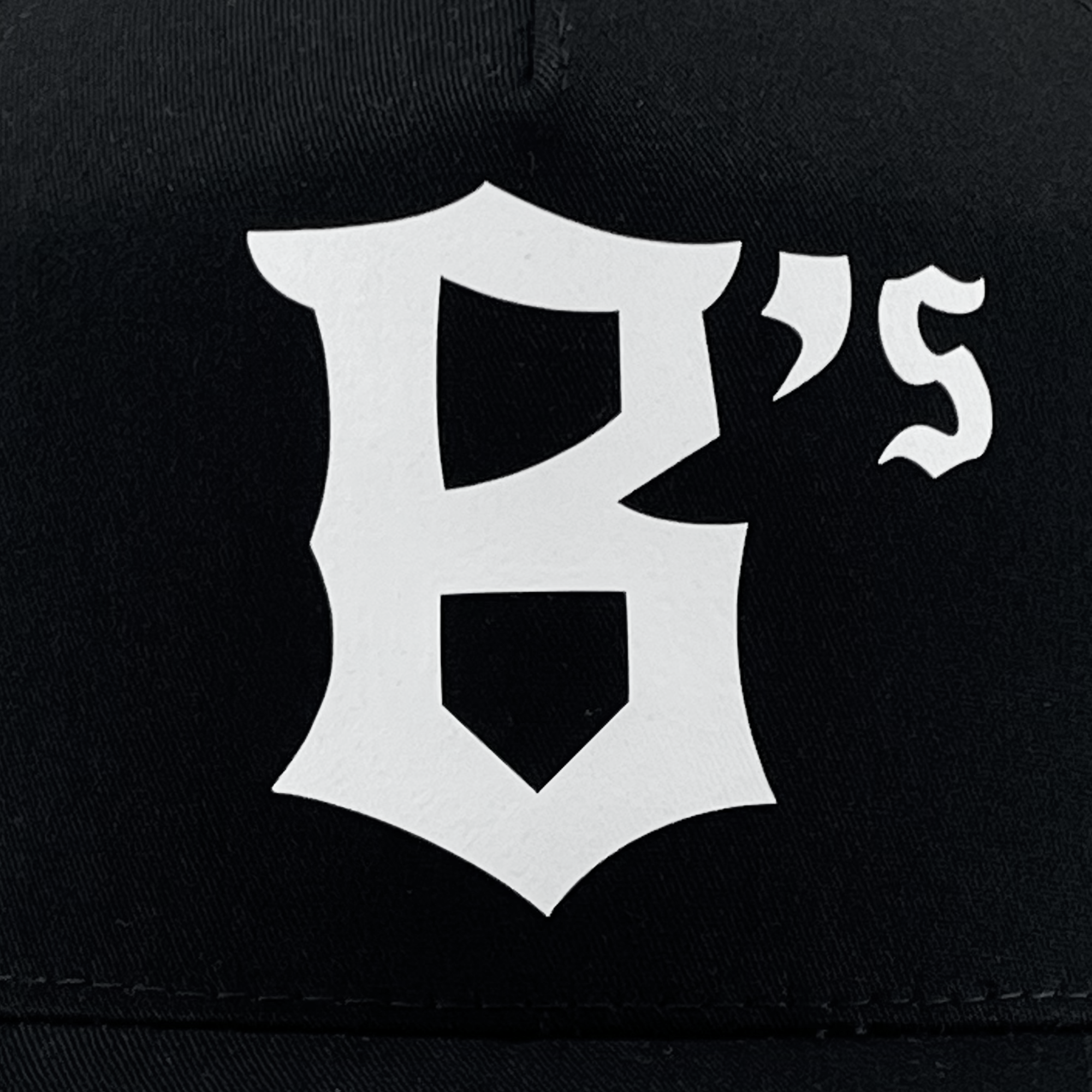 The Oakland Ballers B Trucker is a black cap with a striking white stylized "B" emblem on the front. Next to the “B,” a small white apostrophe and the letter “s” create "B's." This bold design proudly represents the Oakland Ballers, a professional baseball team in the Pioneer League.