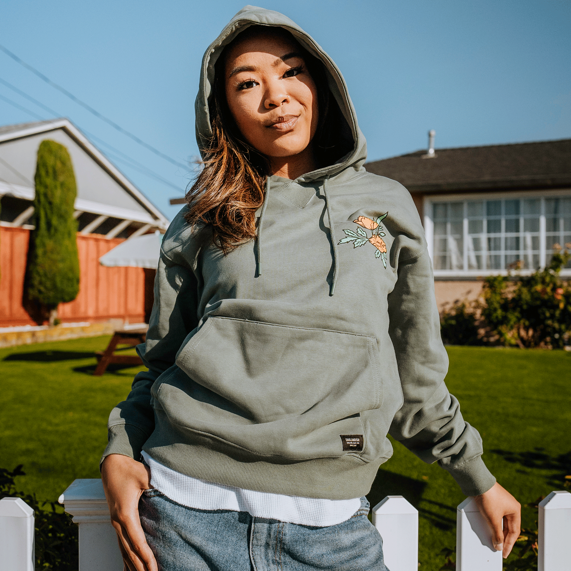 A person stands outdoors, wearing an Oaklandish Oakland Dream Premium Hoodie with a floral design on the chest and the hood up. They lean casually on a white fence with one hand in their pocket. There's a green lawn, a wooden fence, and two houses in the sunny background. They have a relaxed expression.