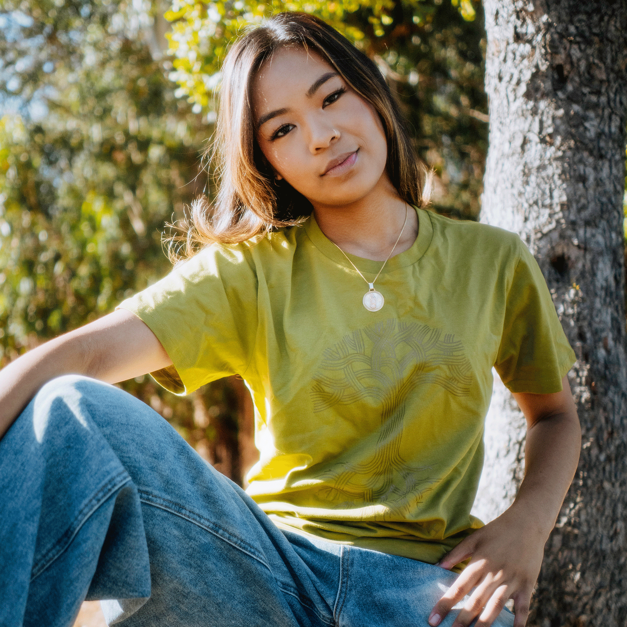 A young woman with long brown hair sits casually outdoors, leaning against a classic tree trunk. She wears an Oaklandish Classic Logo Tee in light green paired with blue jeans. Her relaxed pose and serene expression are highlighted by the sunlight filtering through the tree leaves in the background.