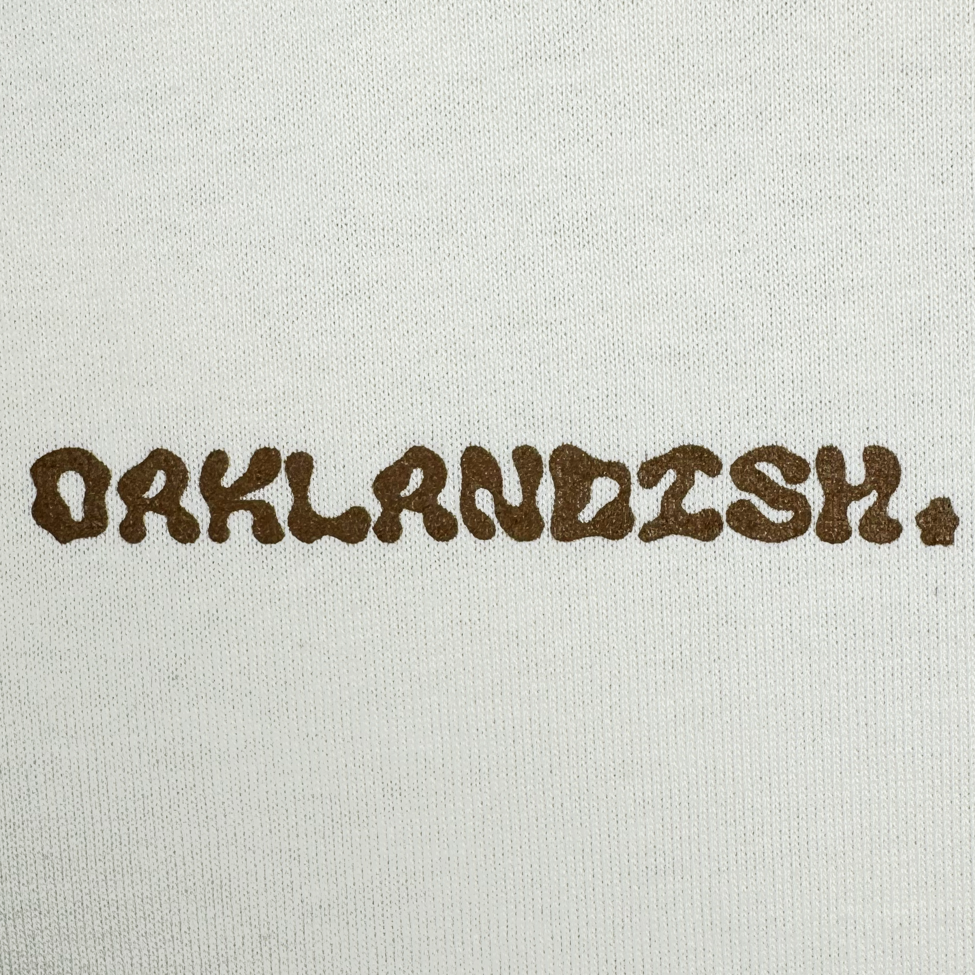 The image displays the word "OAKLANDISH" in textured brown letters on a plain white background, reminiscent of the Grow Your Roots Heavy Tee from Oaklandish, designed for a boxy fit that's popular in men's sizing.