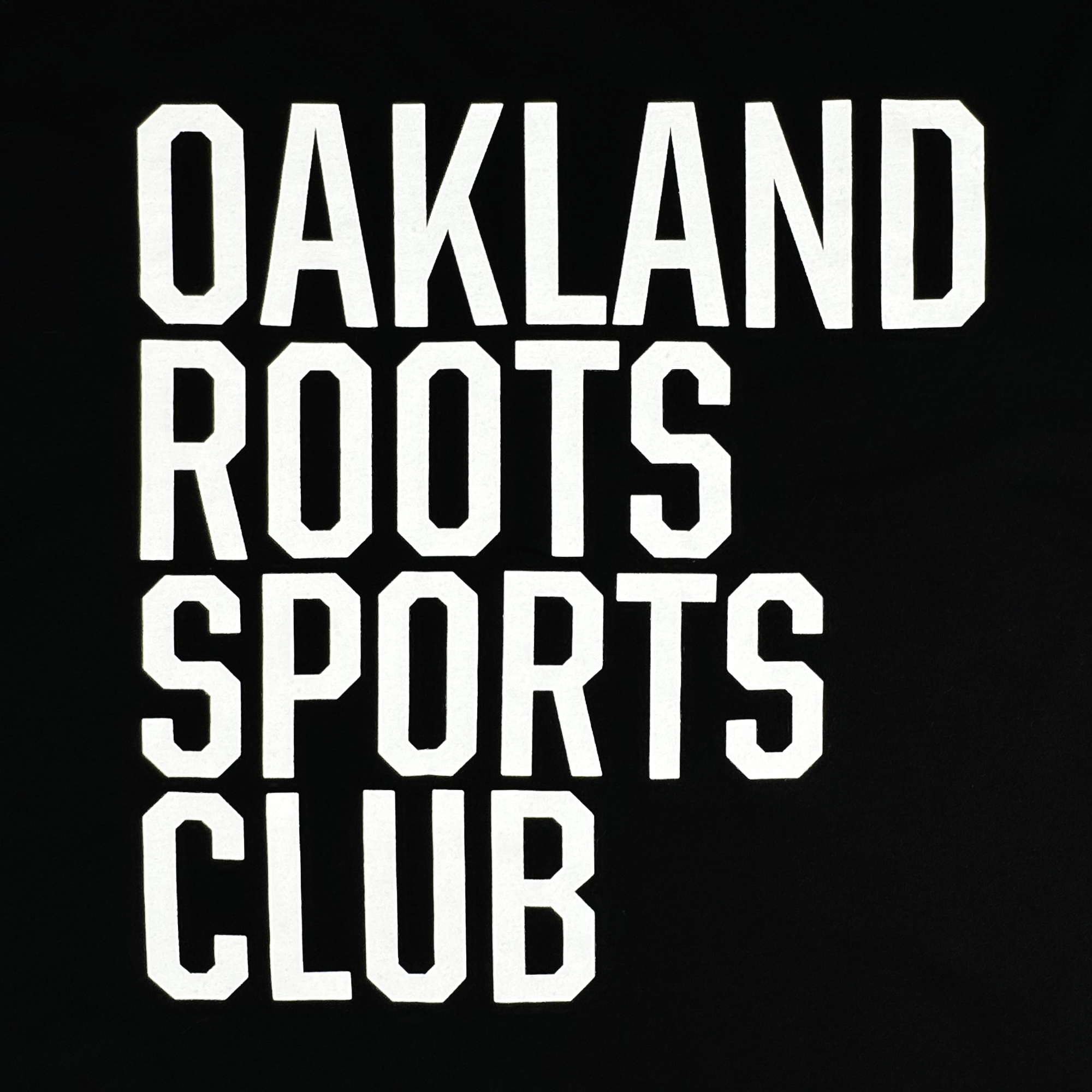 The ORSCtone Tee from Oakland Roots SC features bold white text on a black background with "OAKLAND ROOTS SPORTS CLUB" in a thick, blocky font, embodying the Oaklandish Classic style and highlighting the club’s commitment to social good.