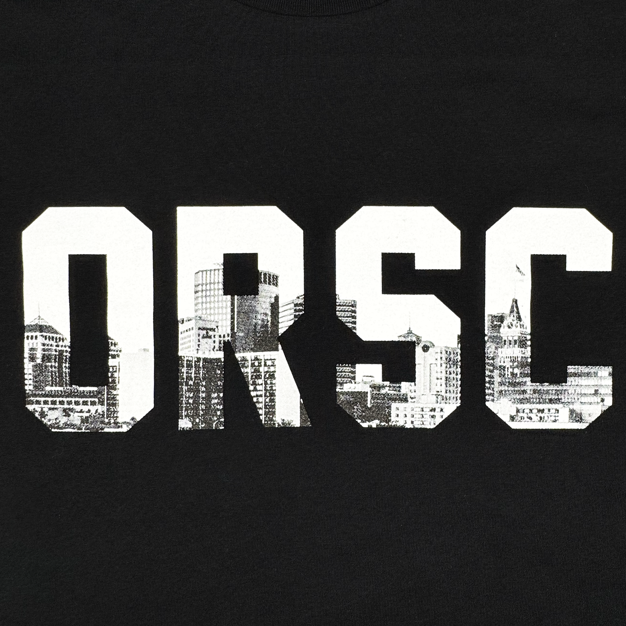 The ORSCtone Tee by Oakland Roots SC features a black background with bold white "ORSC" letters, containing a detailed black-and-white city skyline of buildings and a pointed tower, symbolizing the brand's dedication to social good through urban artistry.