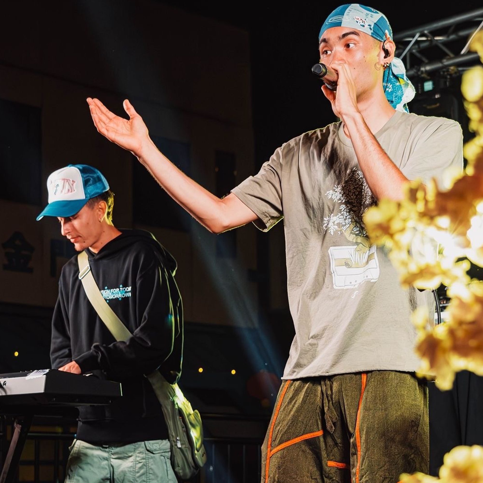 Two musicians perform on stage. The person on the left wears a blue and white cap and black hoodie, playing a keyboard. The person on the right, a Japanese-American rapper named Seiji Oda, holds a microphone while wearing an Oaklandish light-grey Out the Trunk Bonsai Tee, colorful bandana, blue and white cap, and brown pants with orange accents.