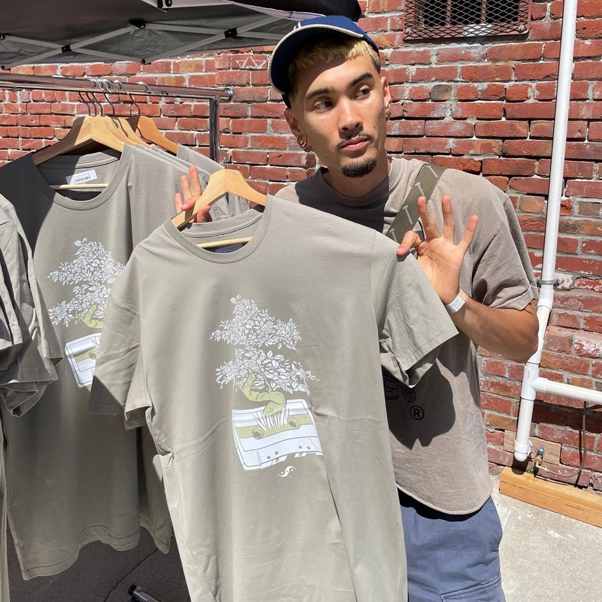 A man stands outside in front of a brick wall, holding an Out the Trunk Bonsai Tee from Oaklandish, featuring a graphic of plants growing out of a cassette tape. He wears a baseball cap, an identical beige tee, and has light facial hair. Nearby, more Out the Trunk Bonsai Tees hang on a rack next to him, capturing the unique style reminiscent of Seiji Oda from Oakland.