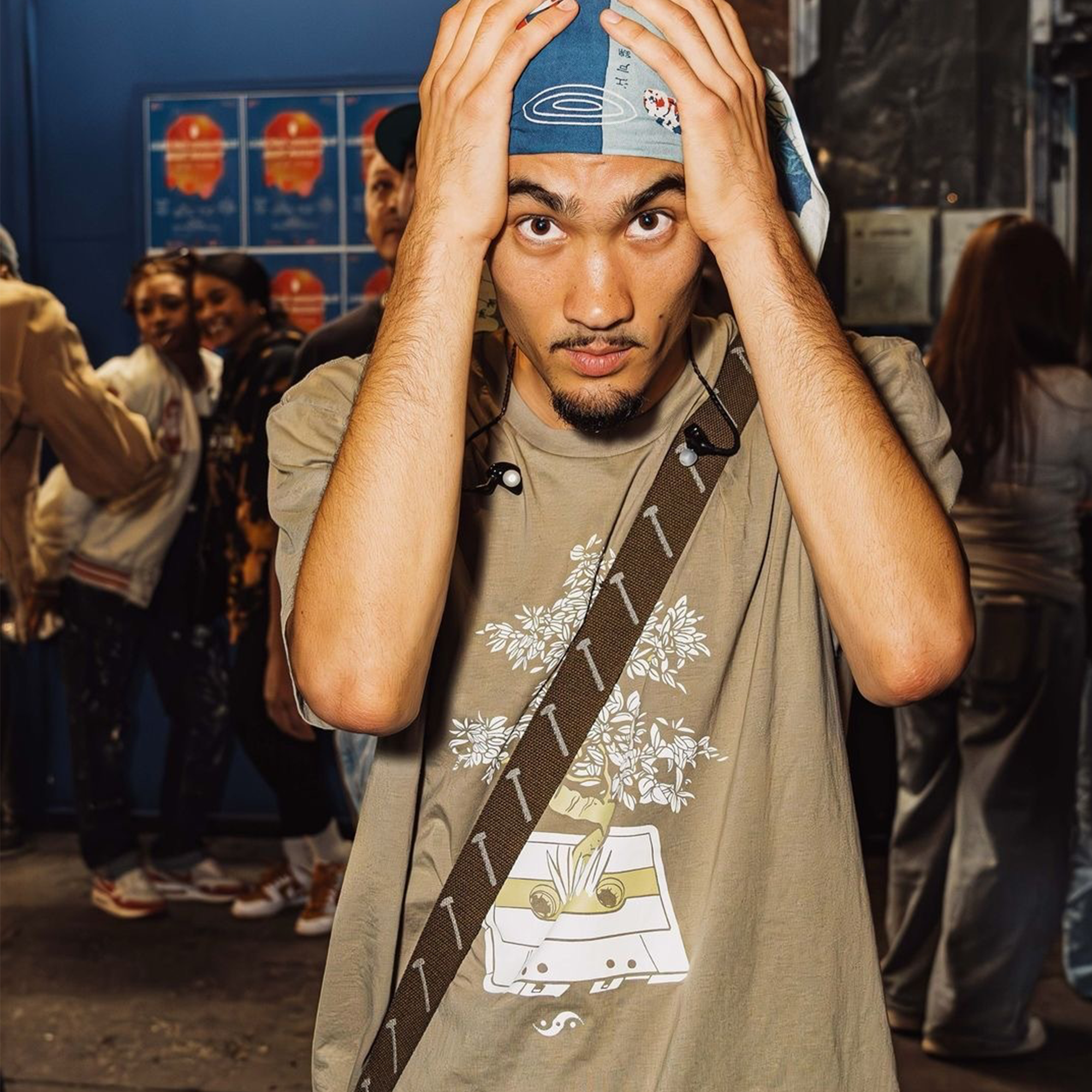 A man with a blue headscarf and goatee, identified as Japanese-American rapper Seiji Oda from Oakland, holds his head with both hands. He’s wearing the beige "Out the Trunk Bonsai Tee" featuring a cassette tape and floral design by Oaklandish, along with a strap across his shoulder. He looks directly at the camera with a focused expression, while people in casual attire populate the blurred background.