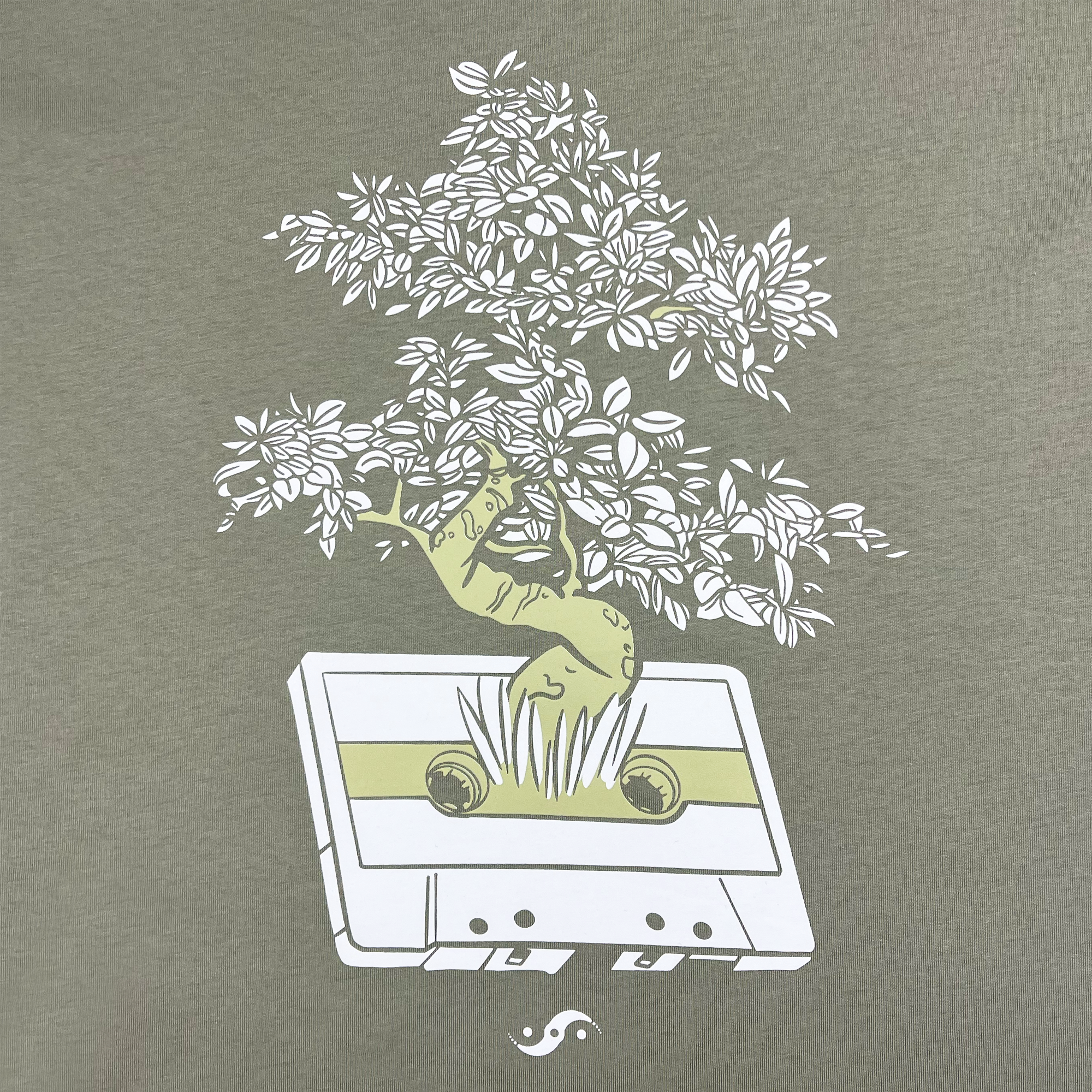 A depiction from Oaklandish's "Out the Trunk Bonsai Tee" features a green cassette tape set against a grey backdrop, sprouting a tree with lush, white leaves. A green lizard with a distinctive brown stripe along its side perches on one of the branches, seamlessly blending into the scene. Beneath the cassette sits a small symbol paying homage to Oakland's Seiji Oda.