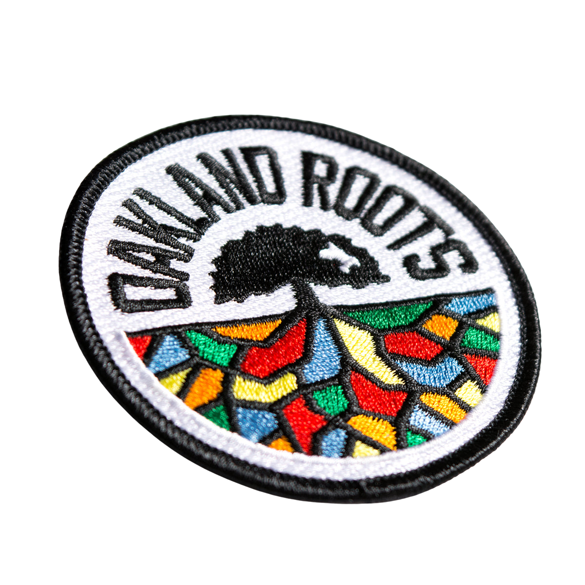 Get your hands on the Oakland Roots SC Patch by Oakland Roots SC, a circular embroidered iron-on patch featuring a black border and "OAKLAND ROOTS" text in black at the top. The center showcases a black tree with roots extending over a vibrant mosaic of red, blue, yellow, and green geometric shapes on a white background—ideal for any Oakland Roots Sports Club fan.