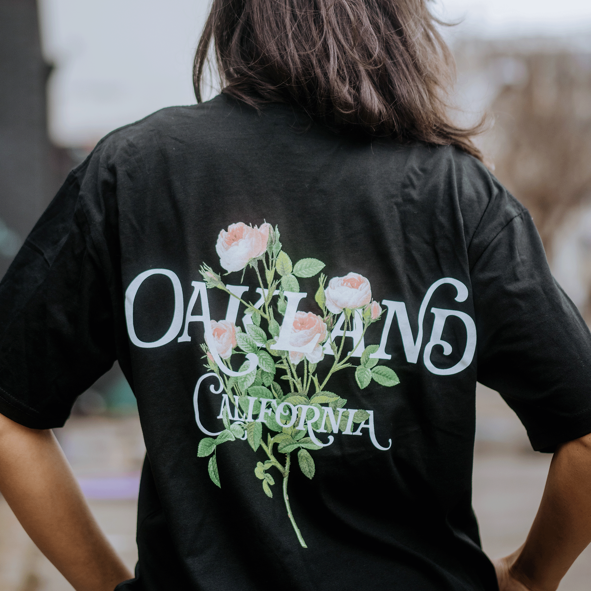 A person with long dark hair wears the Posy Tee by Oaklandish, a classic fit black T-shirt with white and pink roses and "OAKLAND CALIFORNIA" in white on the back. Made from 100% cotton, it stands out stylishly against the blurred background.