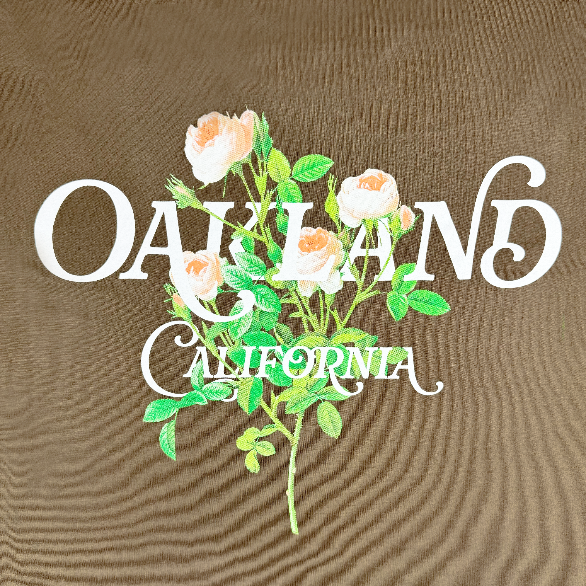 The Women's Posy Tee by Oaklandish features a brown color with bold white "OAKLAND" text, artistically accompanied by the word "California" and a flowering rose branch illustration. Made from 100% cotton, this tee offers comfort and style in women's sizing.