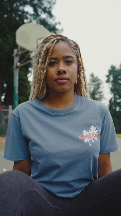 Woman outdoors, sitting on skate deck on a basketball curt wearing this blue tee, while the camera pans around her.