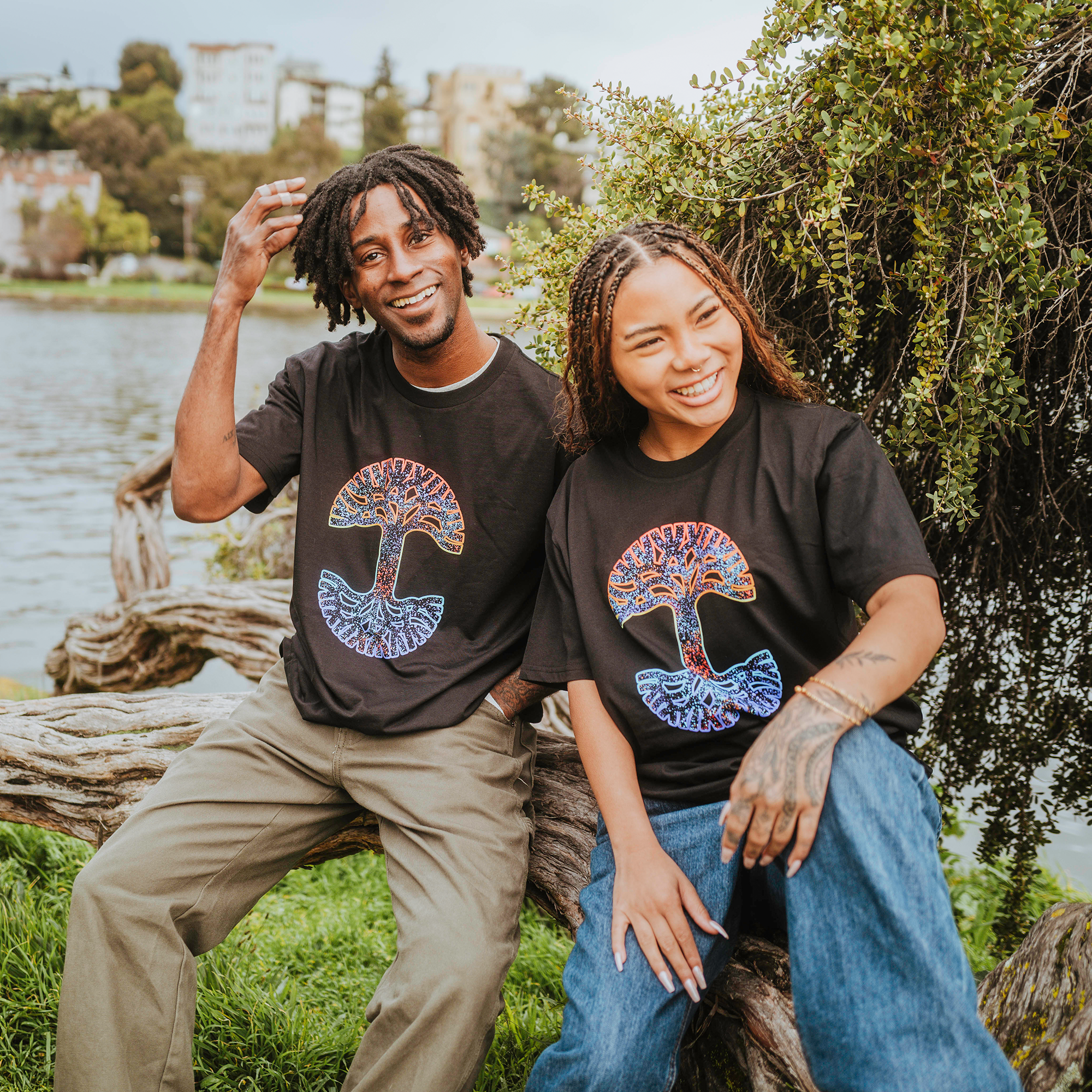 A man and woman smile on a log by a lake, wearing Oaklandish's Prism Tees with colorful mushroom designs. Their 100% cotton shirts harmonize with his short dreadlocks and beige pants, and her braided hair and blue jeans, amidst water, greenery, and buildings under a cloudy sky.