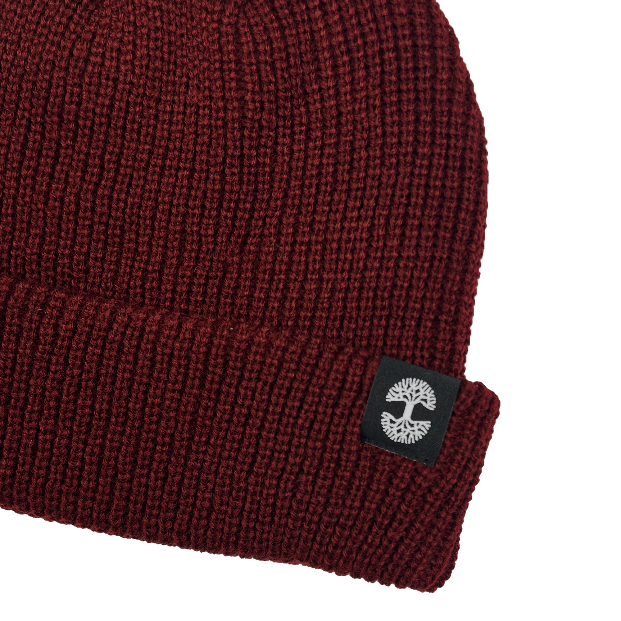 The Ramos Beanie by Oaklandish is displayed. Made from woven acrylic in a maroon shade, it boasts a ribbed texture with wide stitches. A black label with a white tree logo is sewn onto the folded cuff, standing out against the dark maroon of the beanie. The image is set against a plain grey background.