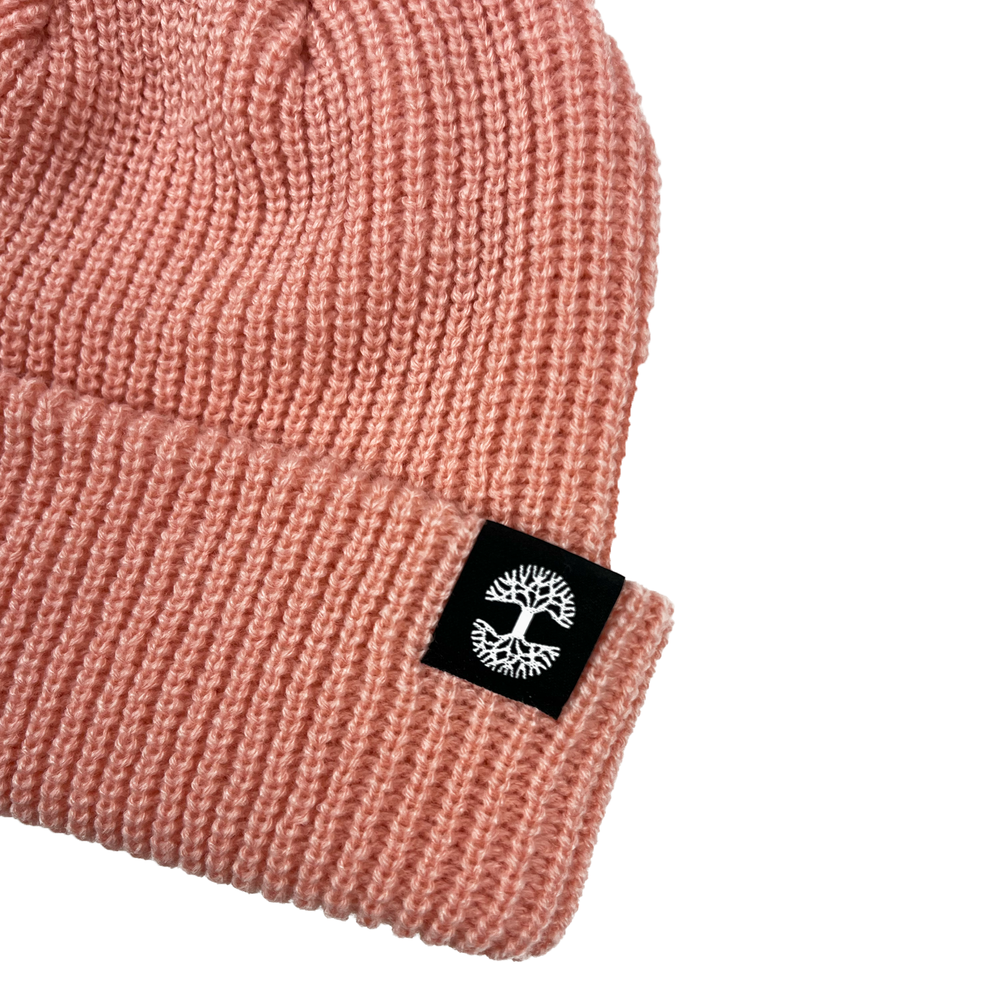 The Oaklandish Ramos Beanie in pink acrylic is depicted with a folded brim, featuring a small black rectangular tag adorned with a white tree logo in the center. The detailed knit texture is prominently visible, enhancing the hat's cozy and warm look. The beanie is presented against a clean white background.