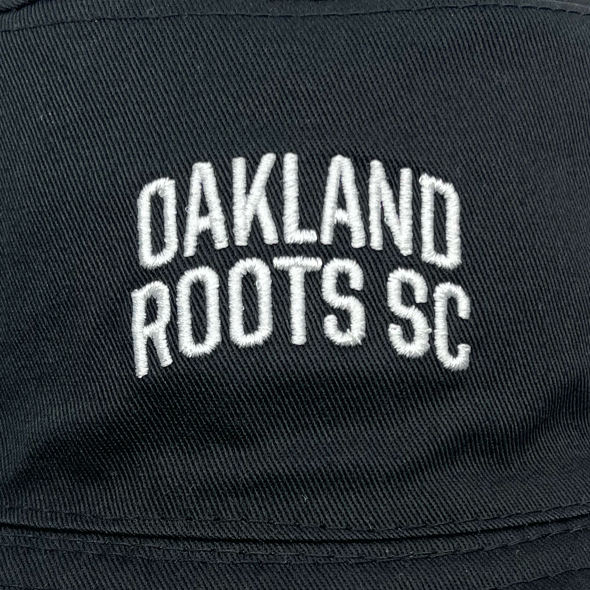 A black fabric surface with white, embroidered text in the center reading "OAKLAND ROOTS SC" in an arched formation. The bold, neatly stitched lettering stands out distinctly against the dark background, suggesting it could be part of the Oakland Soul & Roots SC Reversible Bucket hat by Oakland Roots SC or another stylish piece of their apparel.