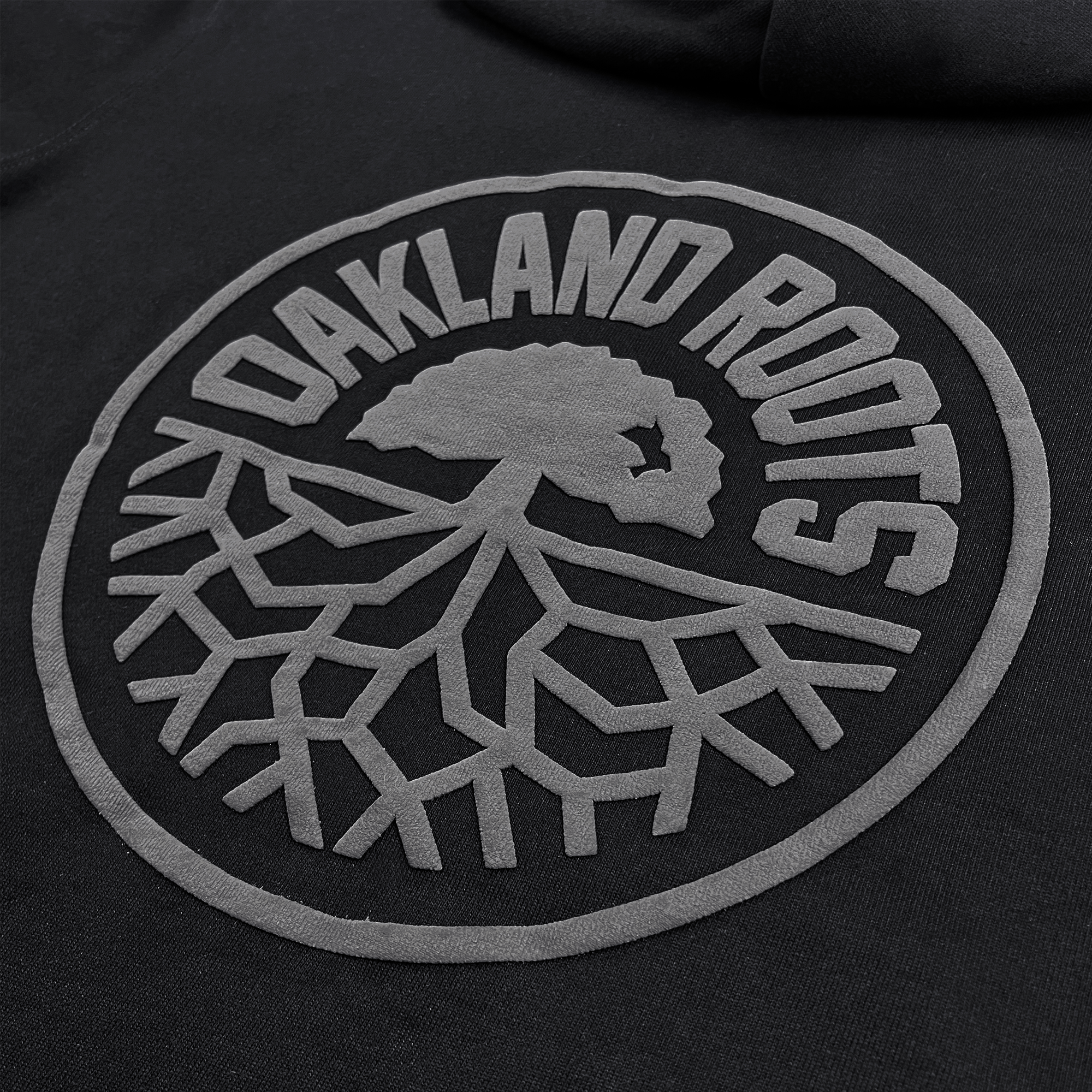 The image shows a close-up of the black organic cotton fabric of the Oakland Roots SC Ascetic Hoodie, displaying the "Oakland Roots" emblem. The emblem features a tree with its roots spreading out below, encapsulated within a circular border. The text "OAKLAND ROOTS" curves around the top half of the circle, all in gray print.