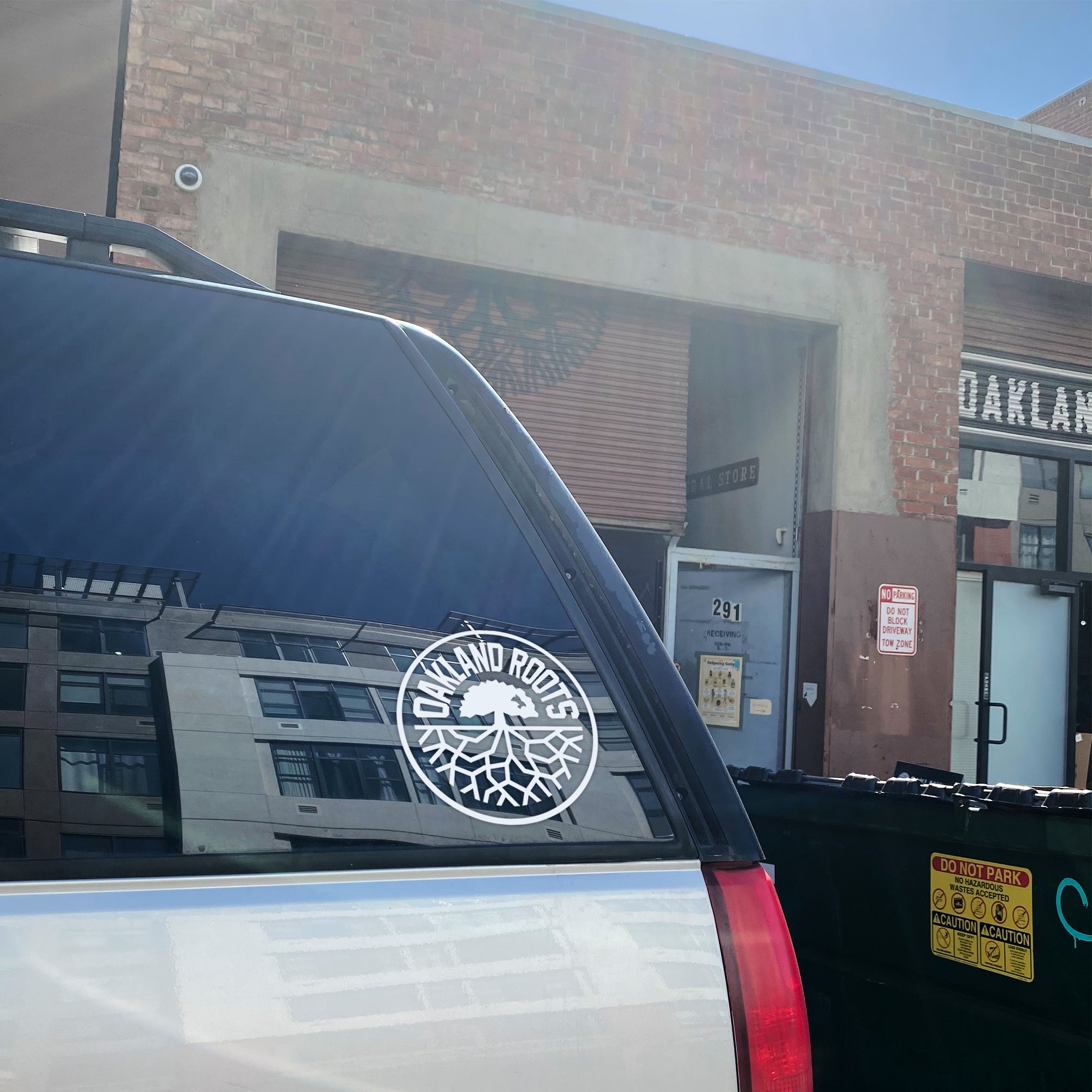 A photo shows the back of a vehicle with an 8" "Oakland Roots SC Decal" on its rear window. The decal showcases tree roots and the text "Oakland Roots," symbolizing the sports club by Oakland Roots SC. The vehicle is parked near a brick building that also features the same "Oakland Roots" text on its facade, with a dumpster visible nearby.