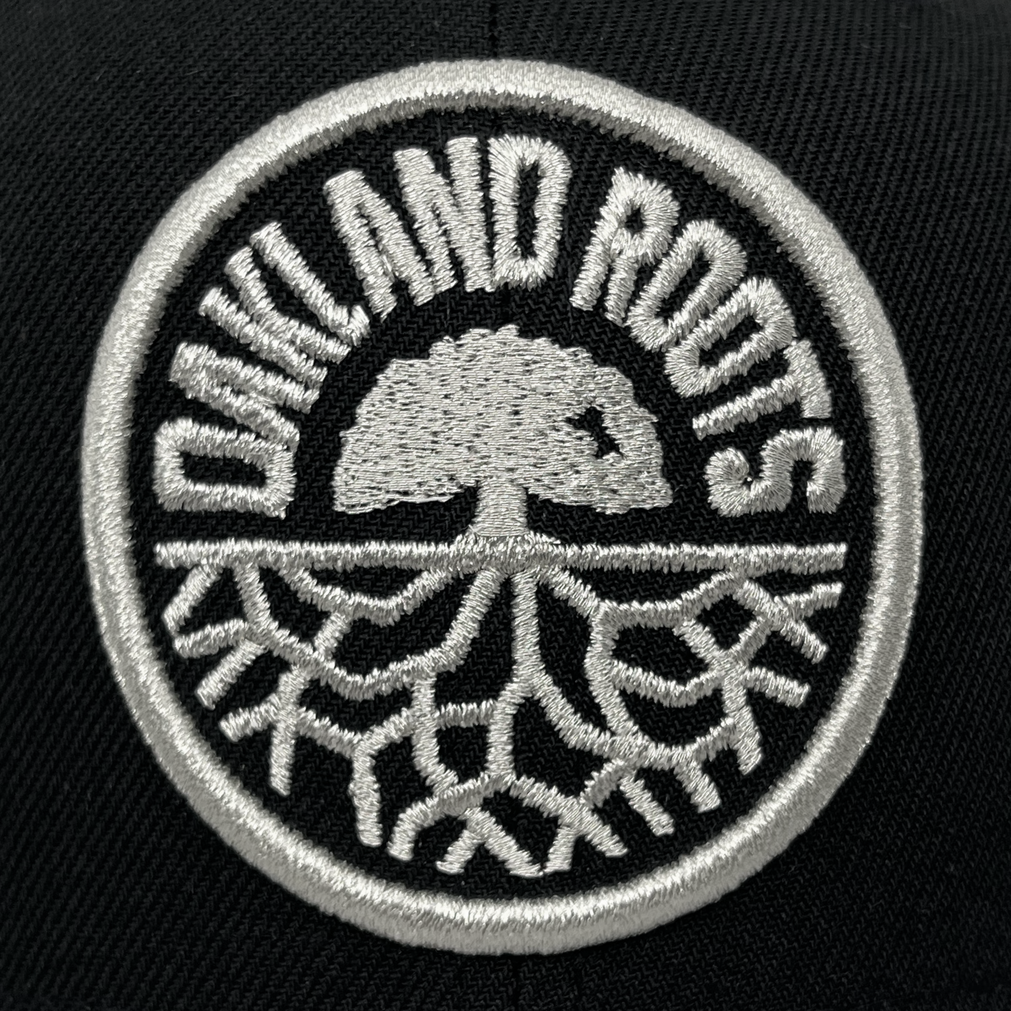 A circular black patch on the fabric of the New Era Oakland Roots SC 59FIFTY Cap showcases the words "Oakland Roots" embroidered in white along the top edge. At its center, a white silhouette of a tree stands over intricate root patterns at the bottom, also in white. The elements are framed by a white embroidered border, making it an ideal accessory to celebrate this vibrant sports club from Oakland Roots SC.